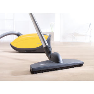 Yellow vacuum cleaner with a silver hose and a vacuuming head on a light wooden floor.