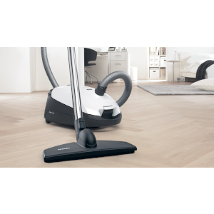 Vacuum cleaner on wooden floor. White body, black head, and a grey hose.