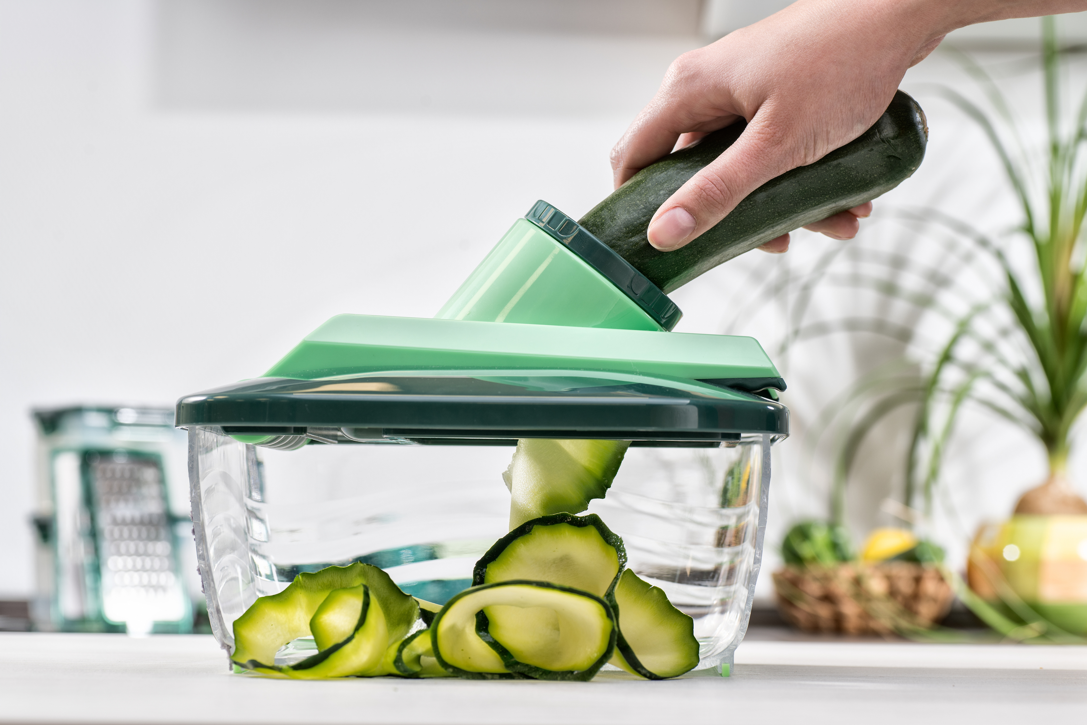 Hand schneidet eine Zucchini mit einem grünen Schneider und Behälter in Streifen.