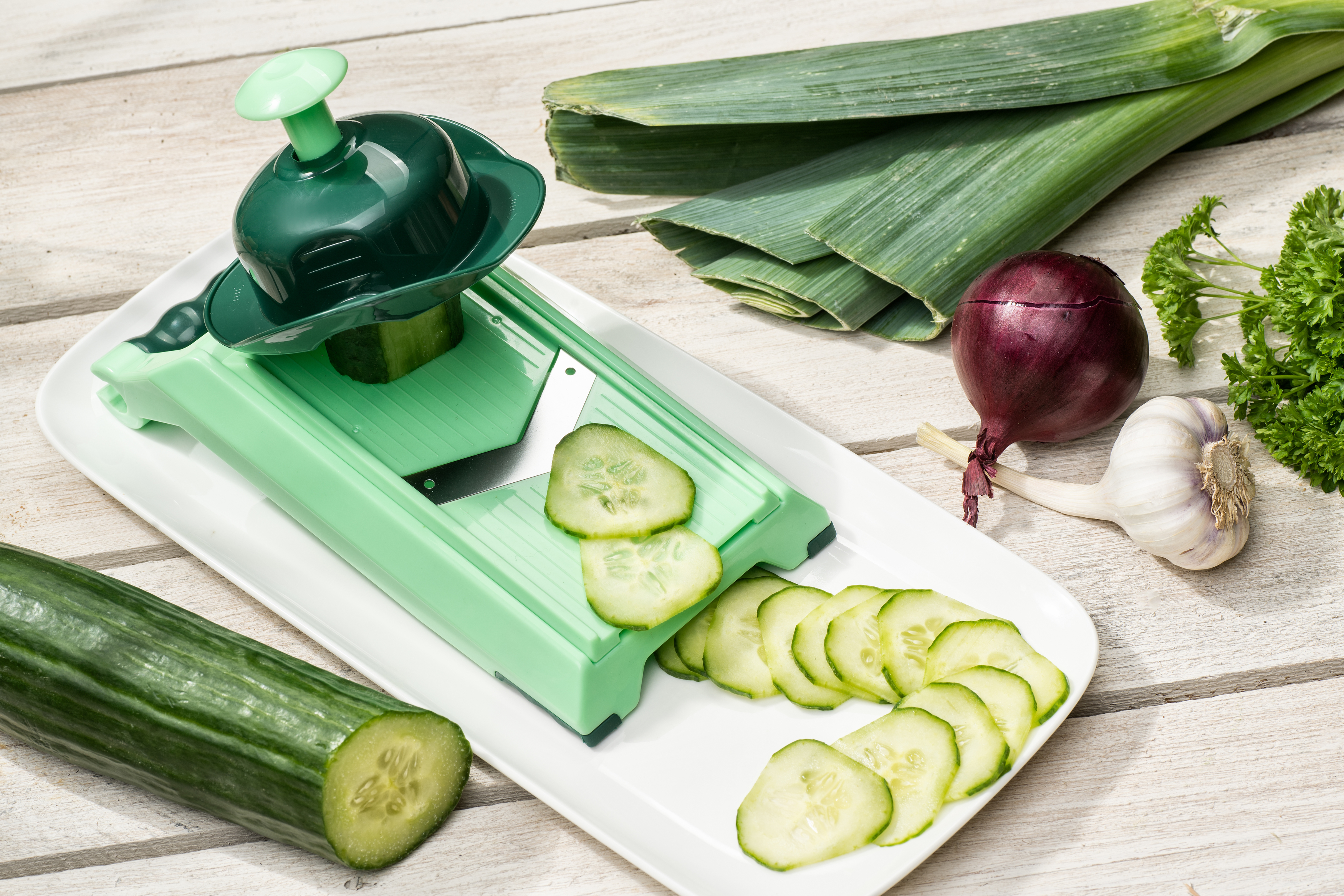 Grüne Mandoline mit Gurkenscheiben. Lauch, Zwiebel, Knoblauch und Petersilie sind in der Nähe.