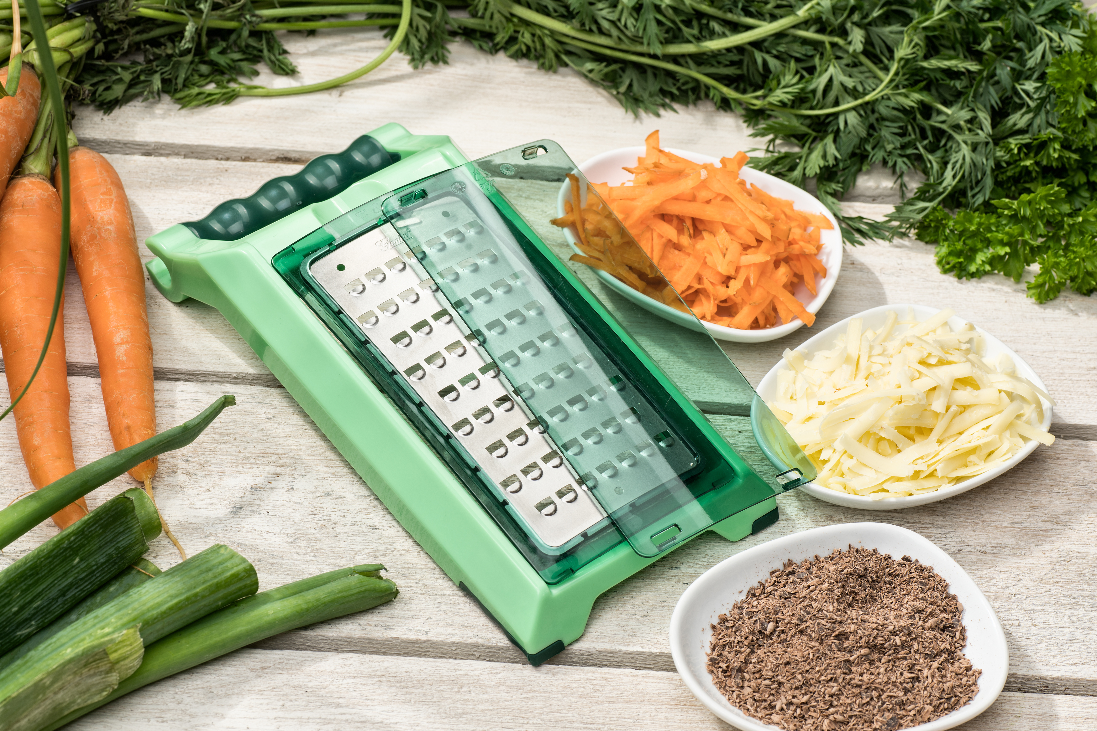 Grüner Reibeisen, Karotten und geriebenes Gemüse auf Holzoberfläche.