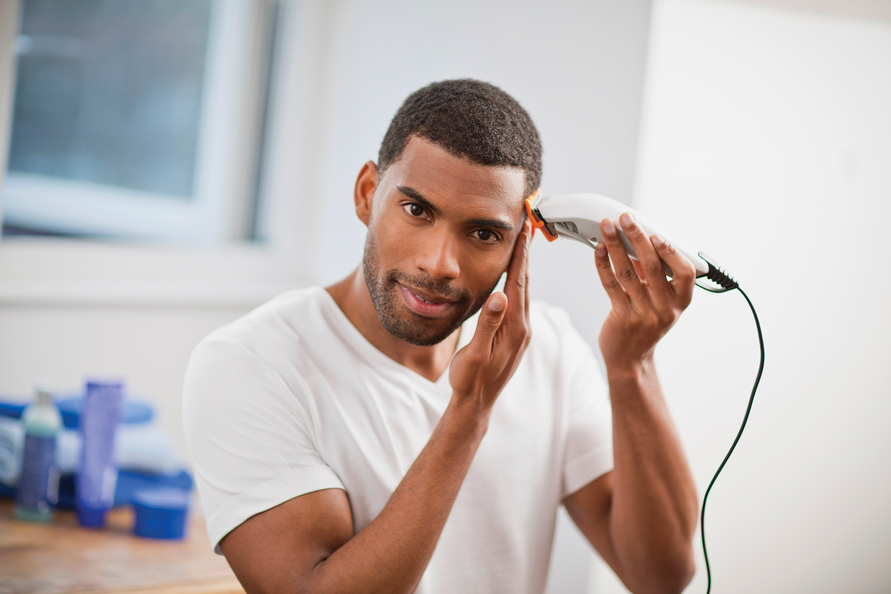 A man is cutting his hair with a trimmer, looking at the camera in a bathroom setting.