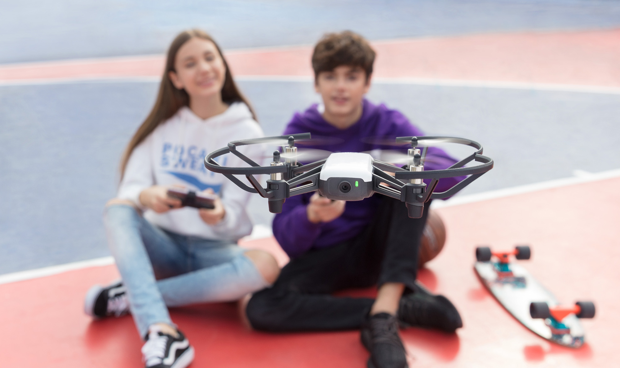 Drone flying over a basketball court. Two teens are sitting on the ground, one holding a controller.
