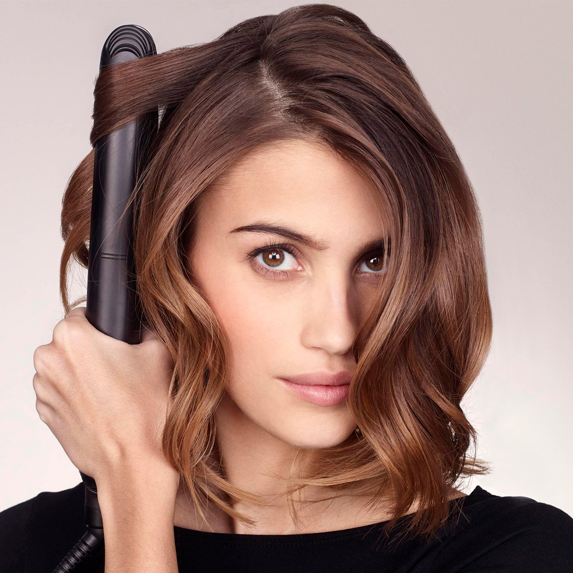 Frau mit braunen Haaren, hält einen Haarglätter. Blick in die Kamera.