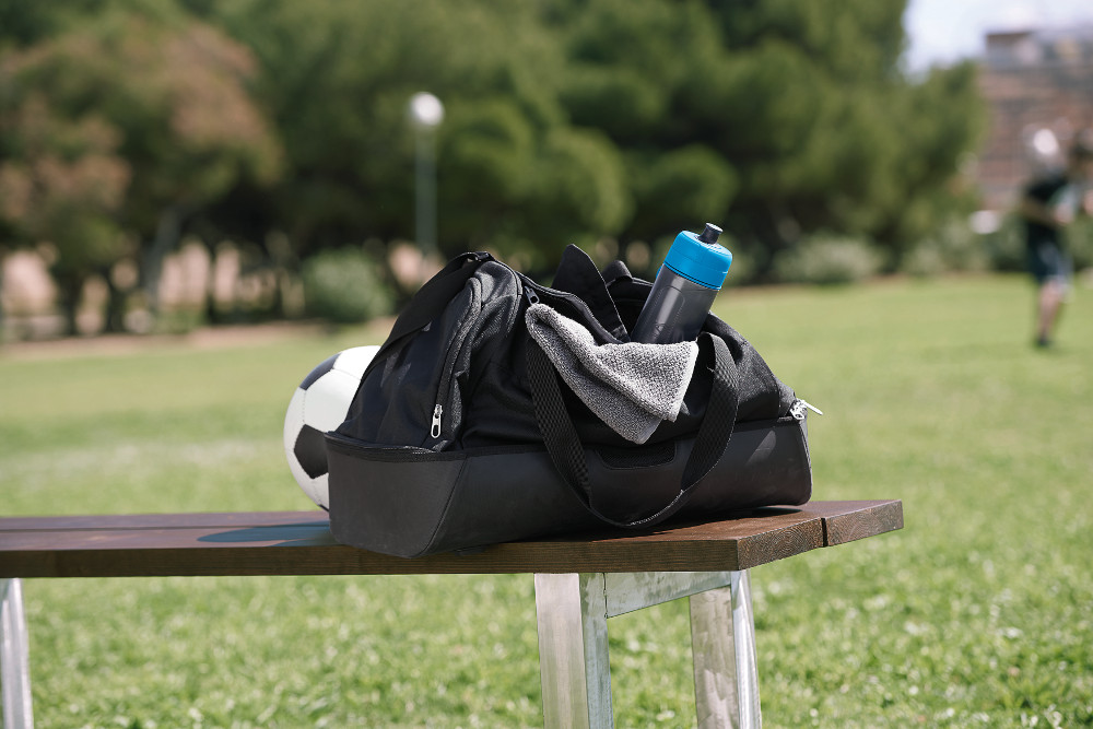 A black sports bag, soccer ball, and water bottle sit on a bench. A person is in the background.