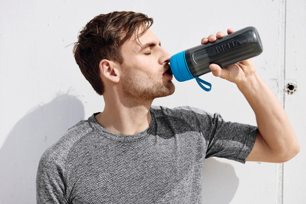 A man drinking from a black and blue Brita water bottle, against a white wall. The man is wearing a grey shirt.