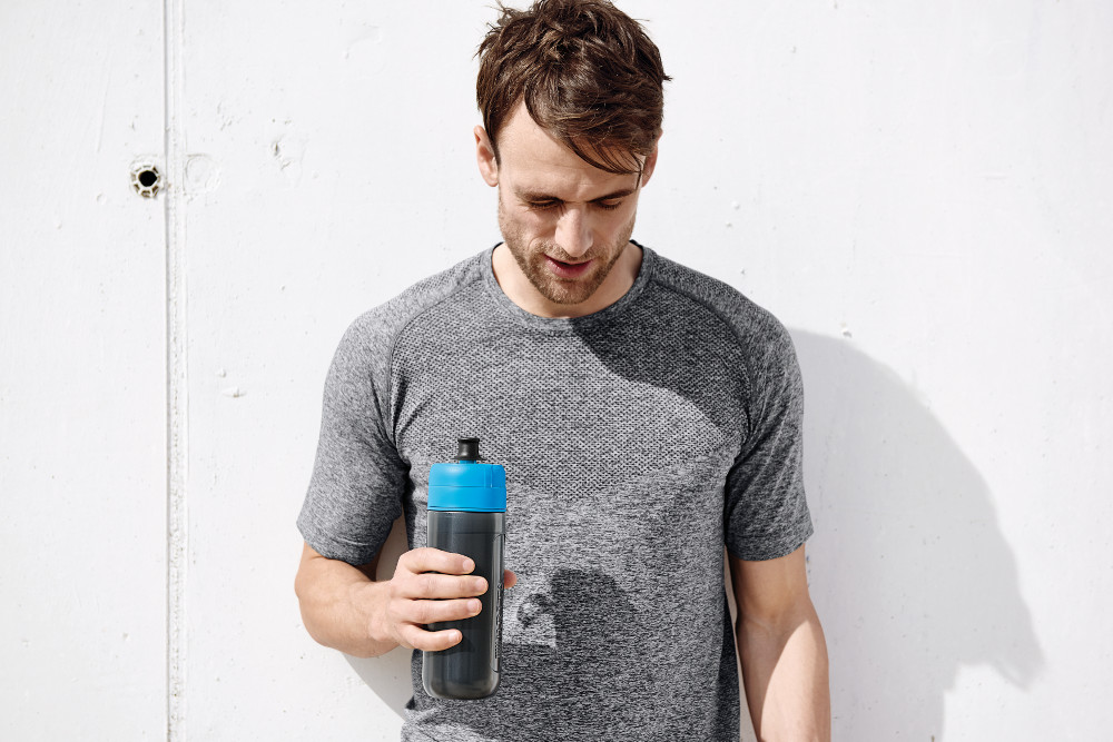 Man holding a water bottle, wearing a grey shirt, standing in front of a wall.