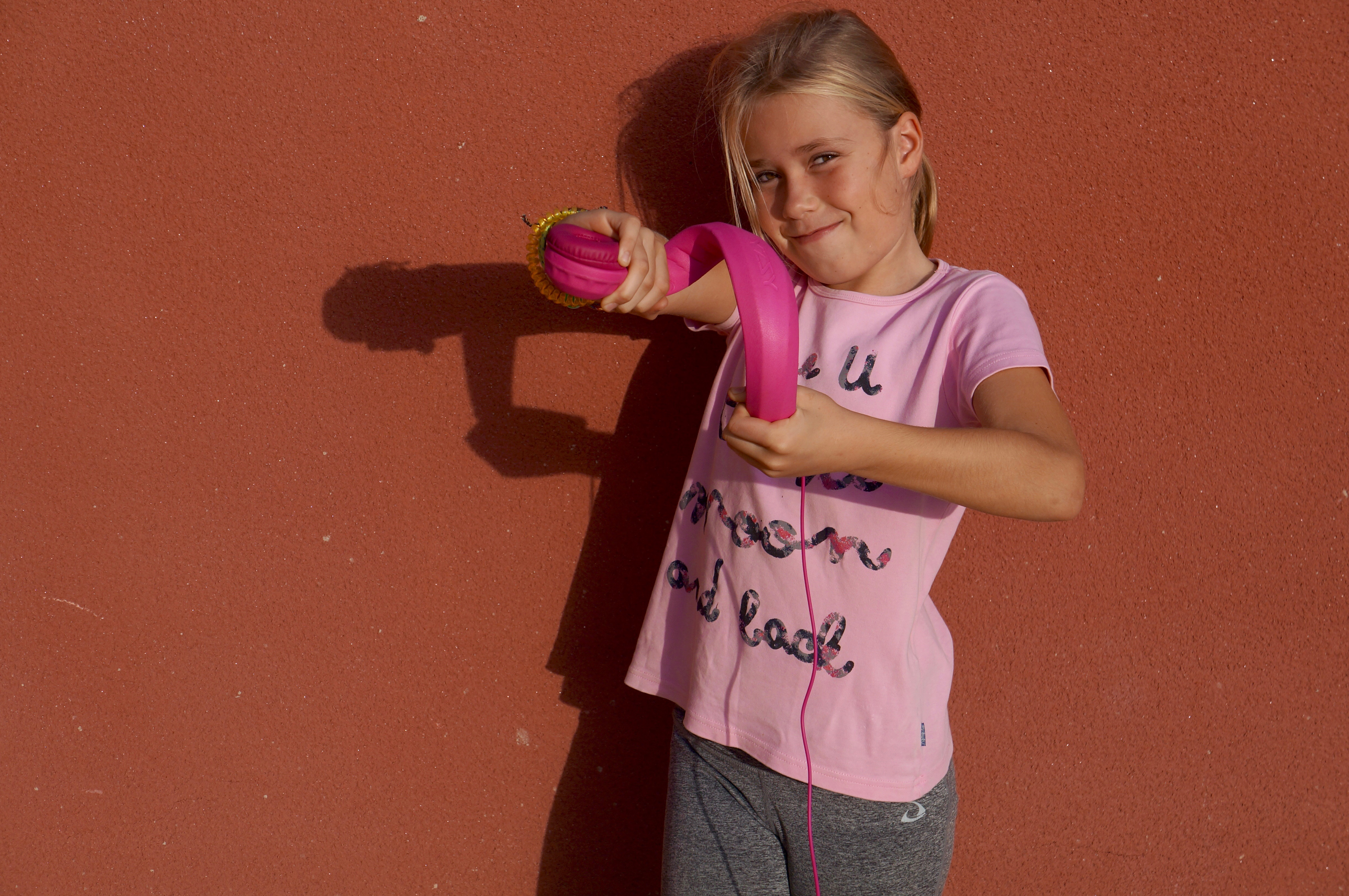 Ein junges Mädchen in rosa Shirt hält pinke Kopfhörer vor roter Wand.