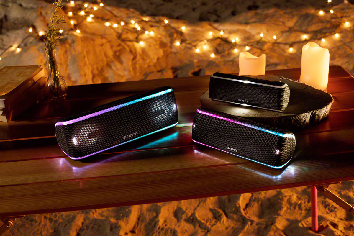 Drei Lautsprecher auf einem Holztisch mit Kerzen und Lichtern im Hintergrund. Die Lautsprecher haben bunte Lichter.