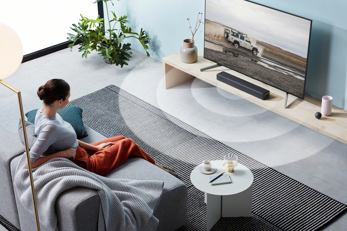 Frau schaut fern mit Soundbar. Lampe, Couchtisch, Sofa und Teppich sind sichtbar.