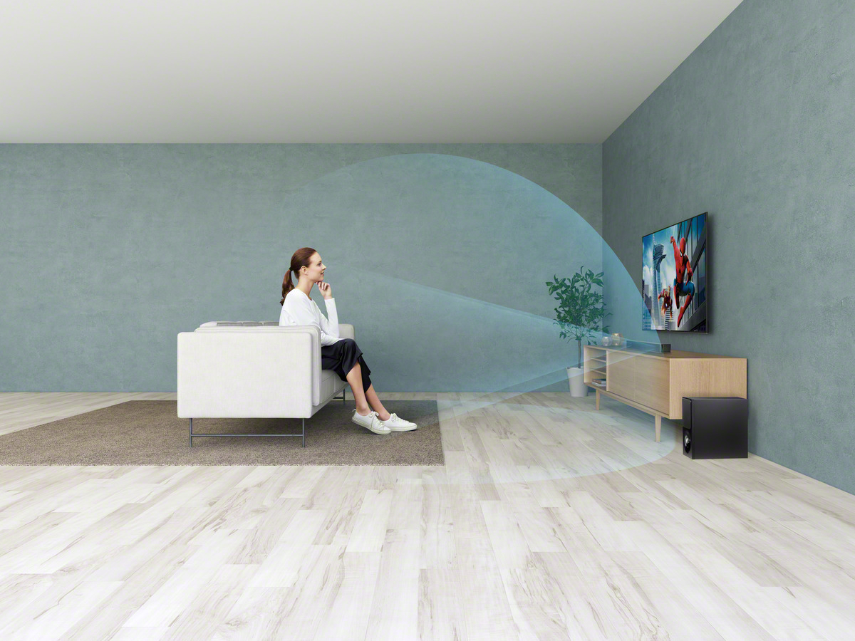 Frau schaut im Wohnzimmer fern. Eine Soundbar und ein Subwoofer sind vorhanden.