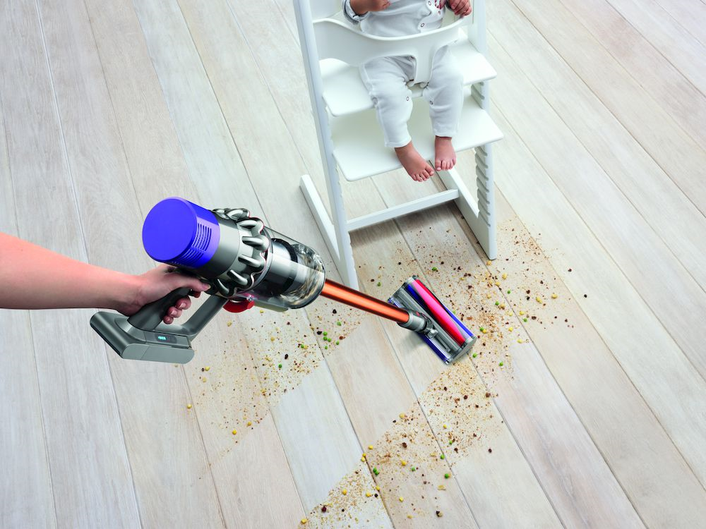 Stofzuiger maakt houten vloer schoon met verspreid voedsel. Een baby zit in een kinderstoel.