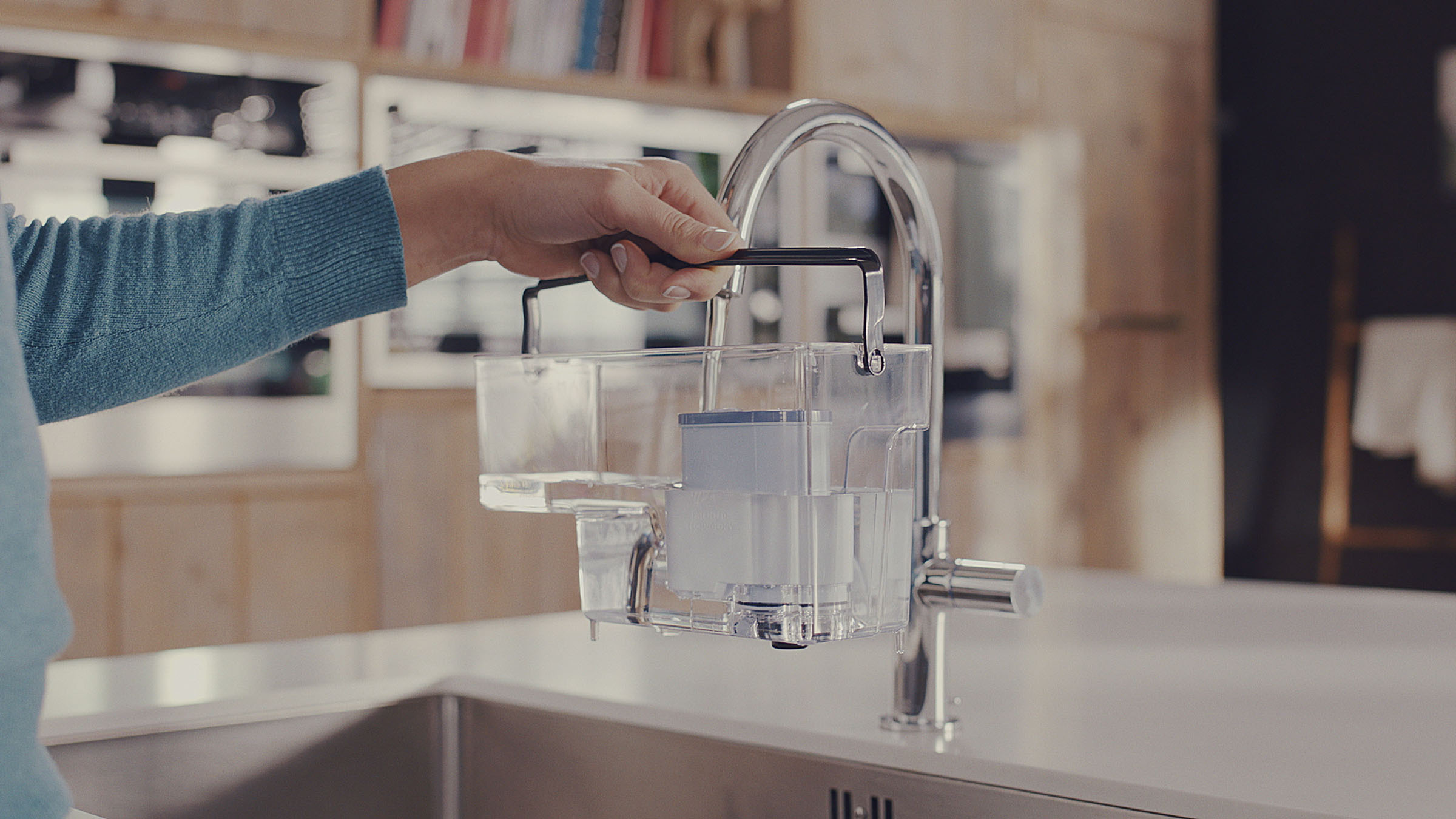 Eine Person hält einen durchsichtigen Behälter mit einem Filter über einem Wasserhahn. Der Wasserhahn ist silber.