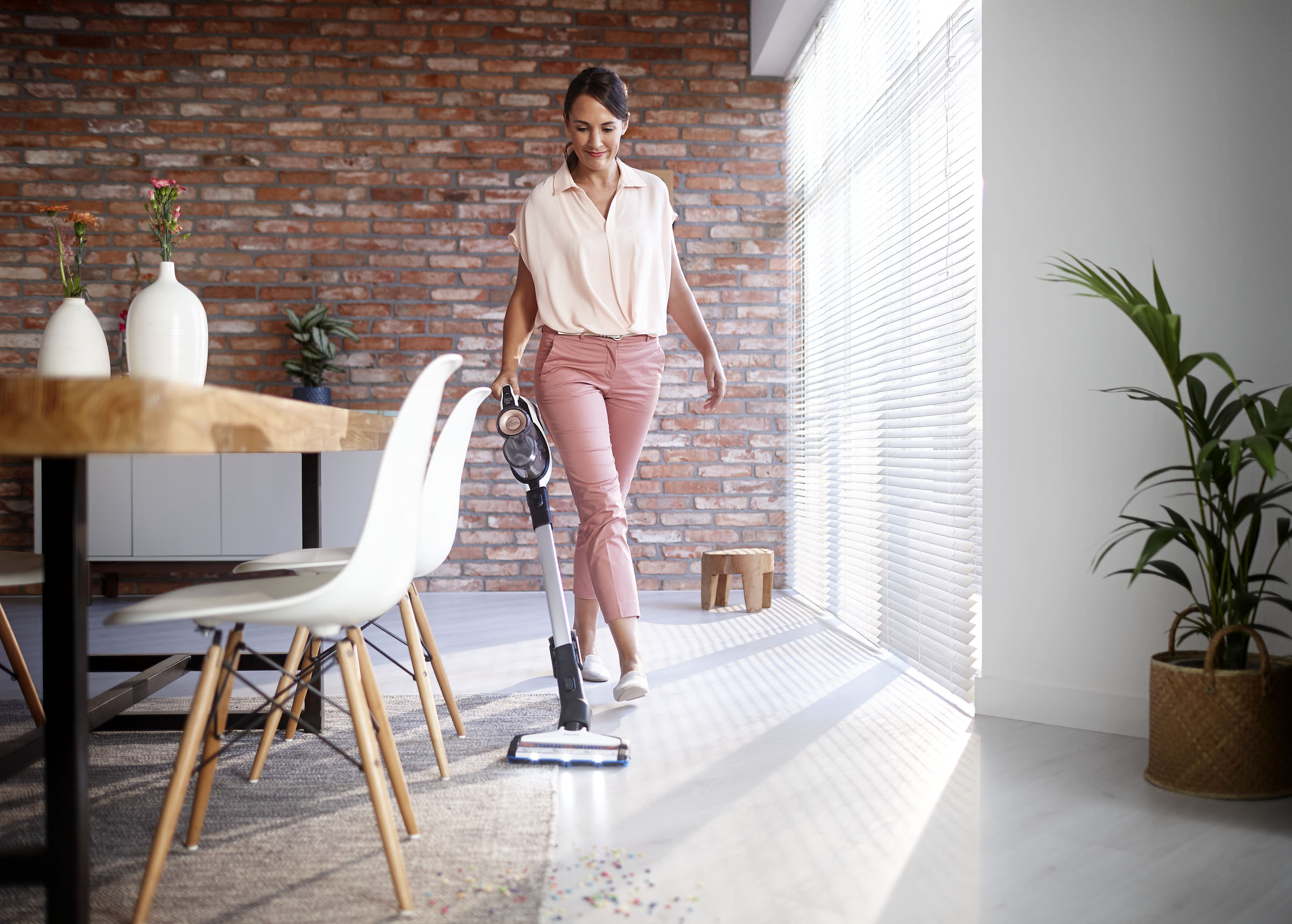 Frau staubsaugt im Wohnzimmer, trägt rosa Hose. Eine Backsteinmauer, weiße Stühle und eine Pflanze sind auch sichtbar.