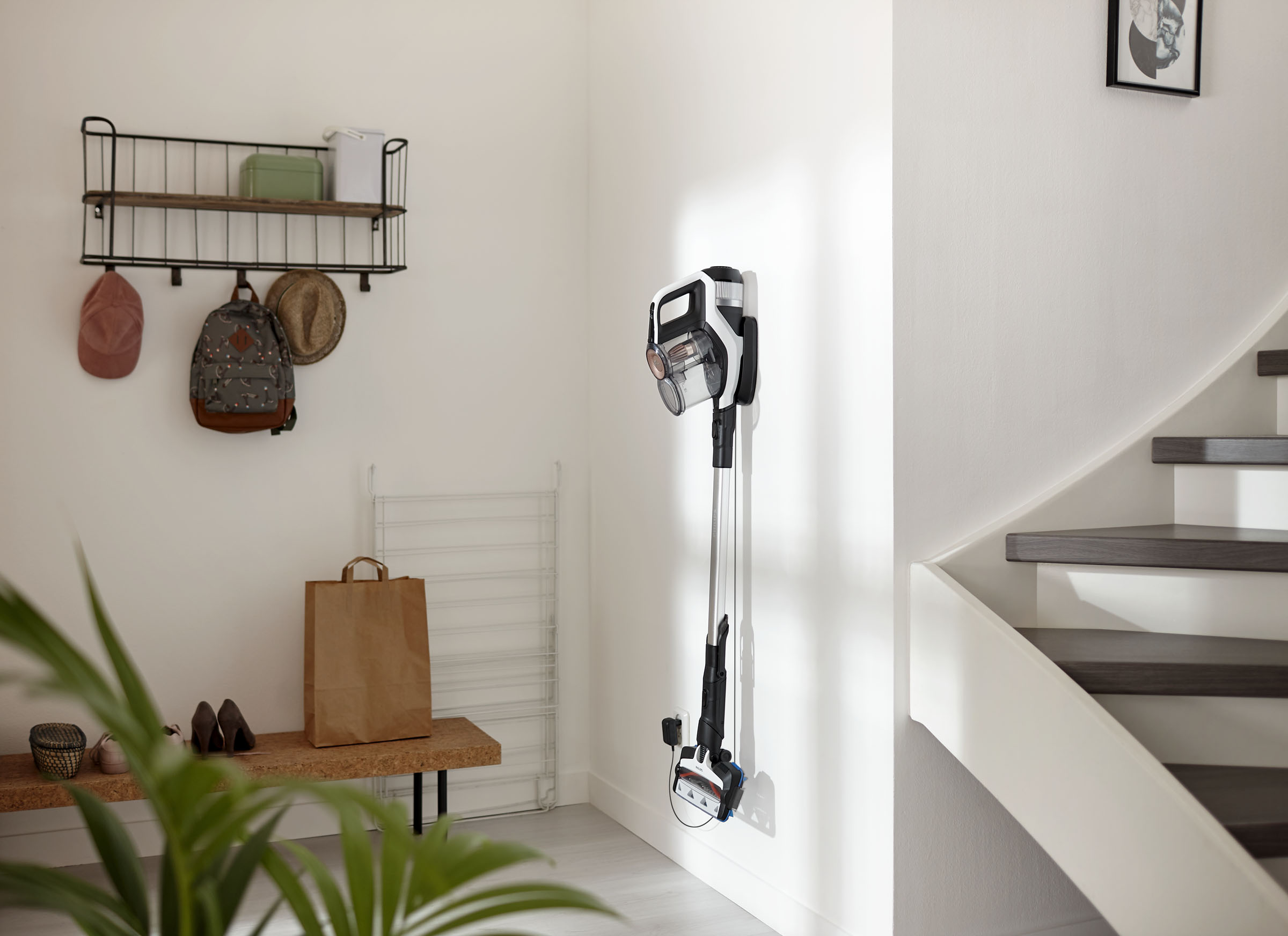 Eine weiße Wand mit Staubsauger, eine Bank mit Schuhen und eine Treppe. Auf einem Regal sind verschiedene Gegenstände.