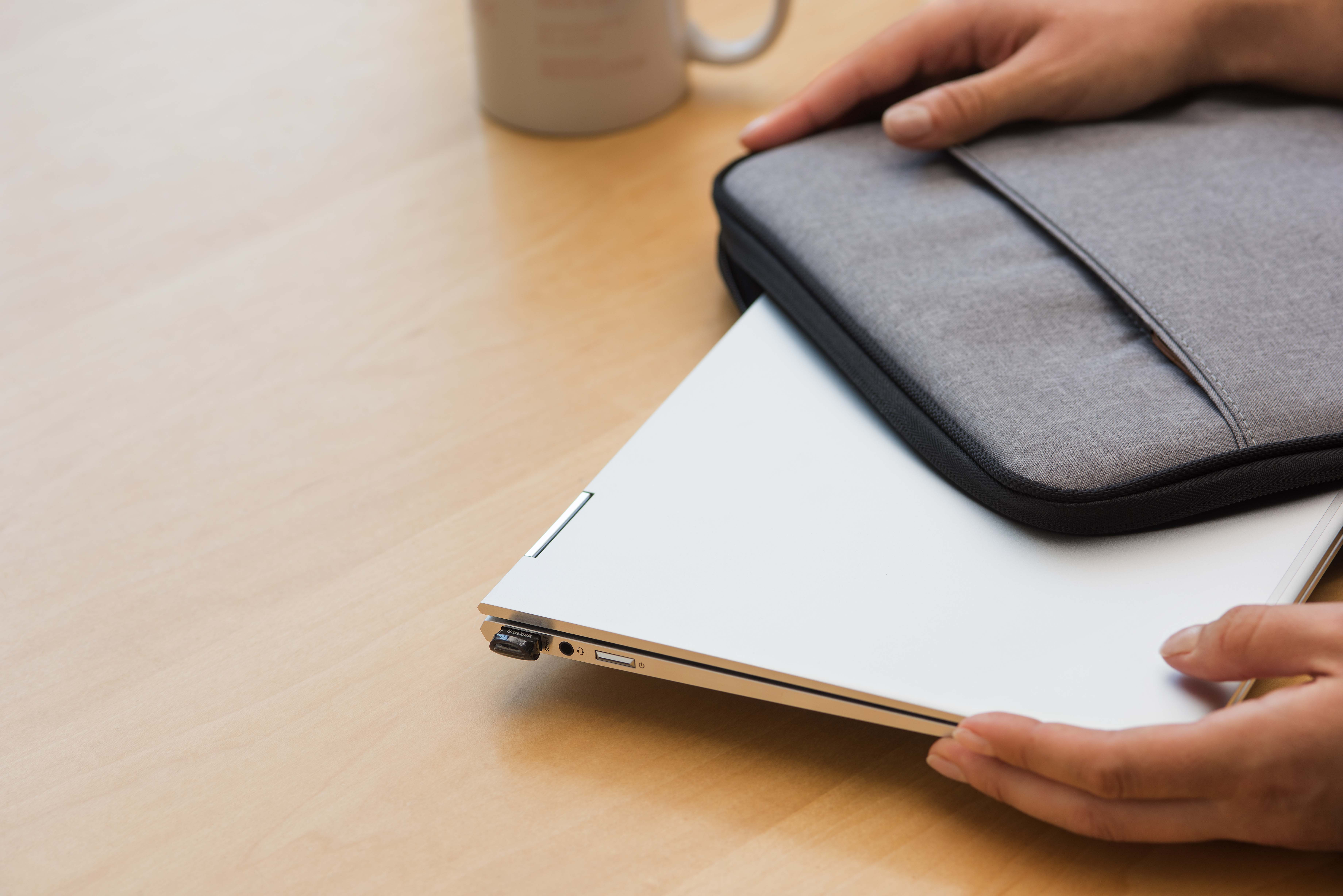 Hands holding a laptop and a gray laptop sleeve on a wooden table. A mug is in the background.