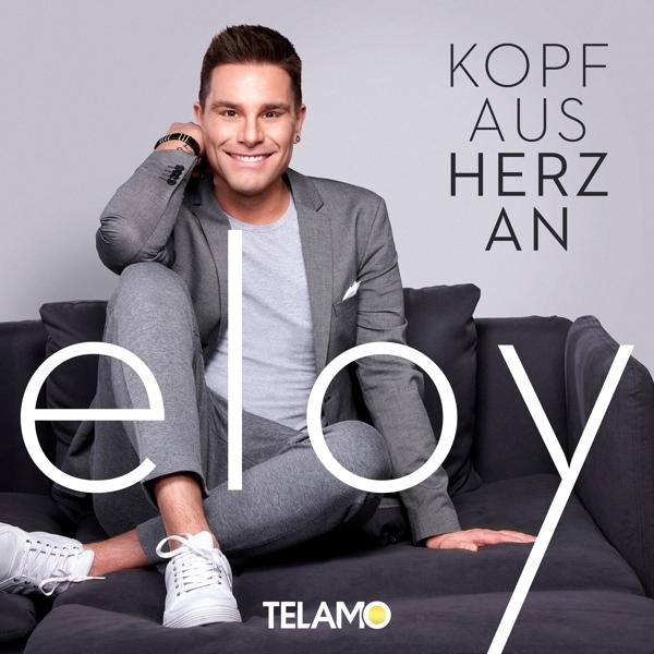 Mann im grauen Anzug sitzt auf einem Sofa und lächelt in die Kamera.