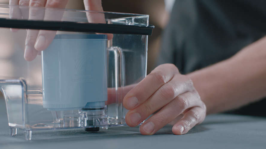 Hände, die einen blauen Filter in einen durchsichtigen Wasserfilterkrug einsetzen.