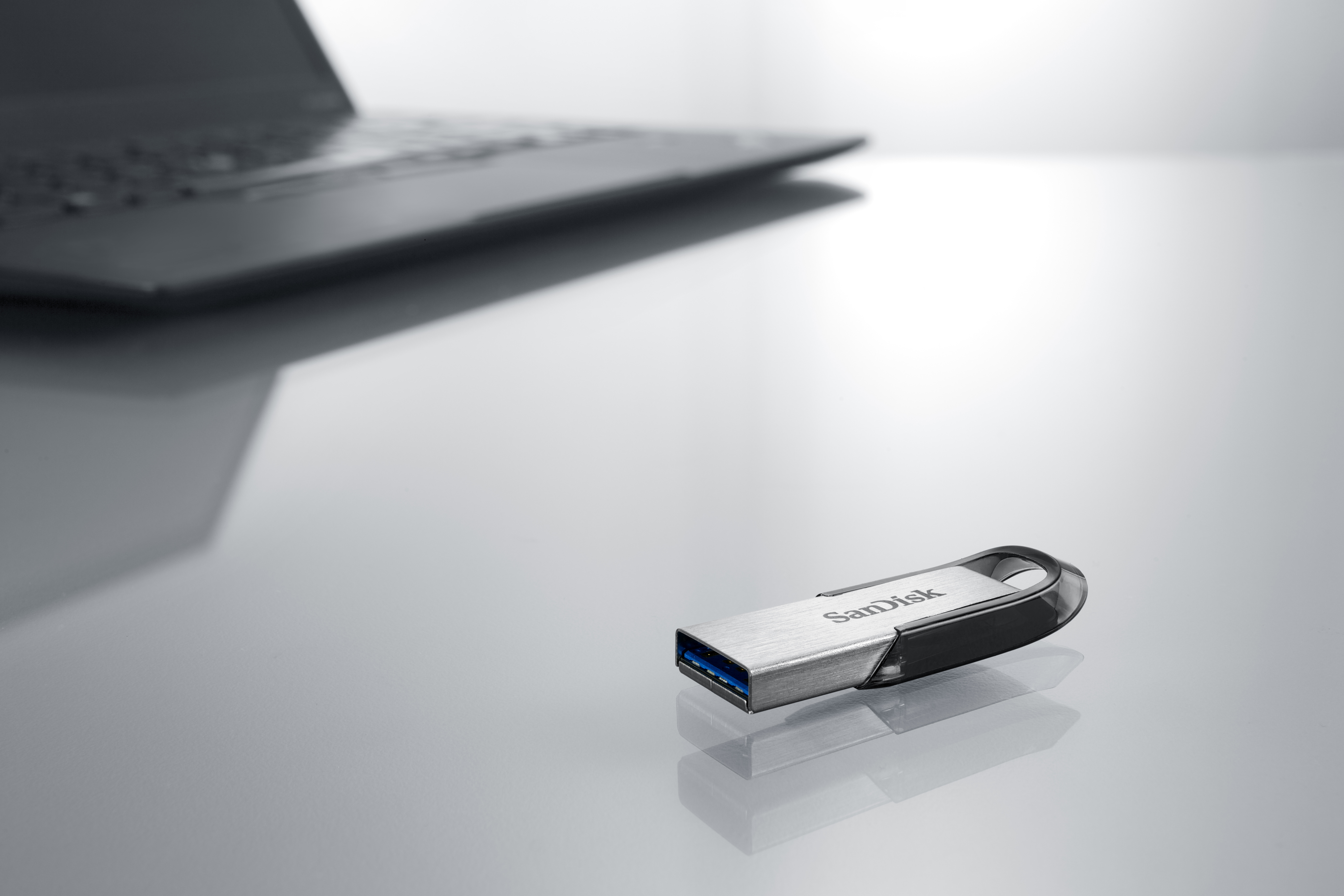 Silver and black SanDisk USB drive on a reflective surface with a laptop in the background.