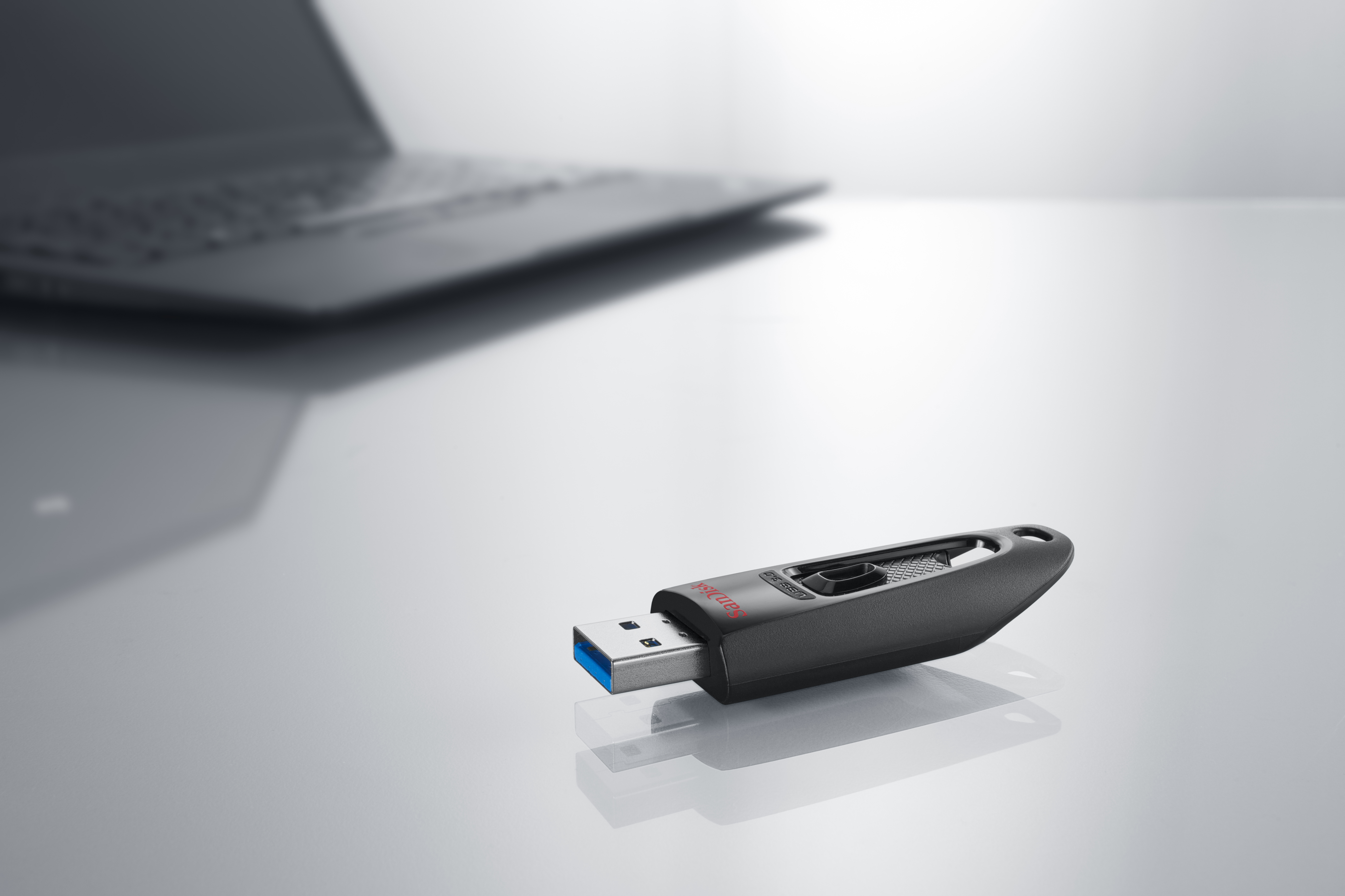 Black SanDisk USB flash drive on a white surface, near a laptop.