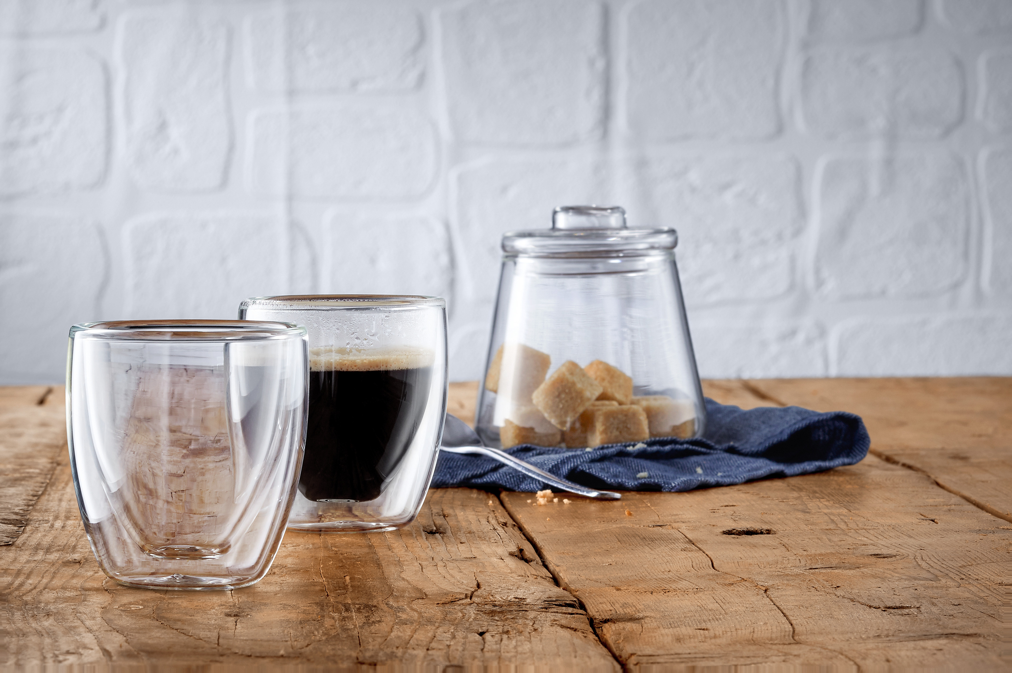 Zwei Gläser mit Kaffee, ein Glas mit Zuckerwürfeln, auf einem Holztisch.