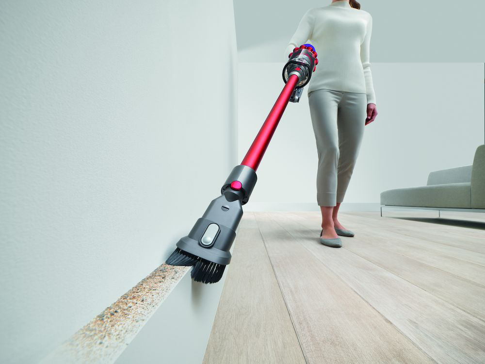Vrouw gebruikt een rode en zilveren Dyson stofzuiger om plint te reinigen. Houten vloer en witte muren.