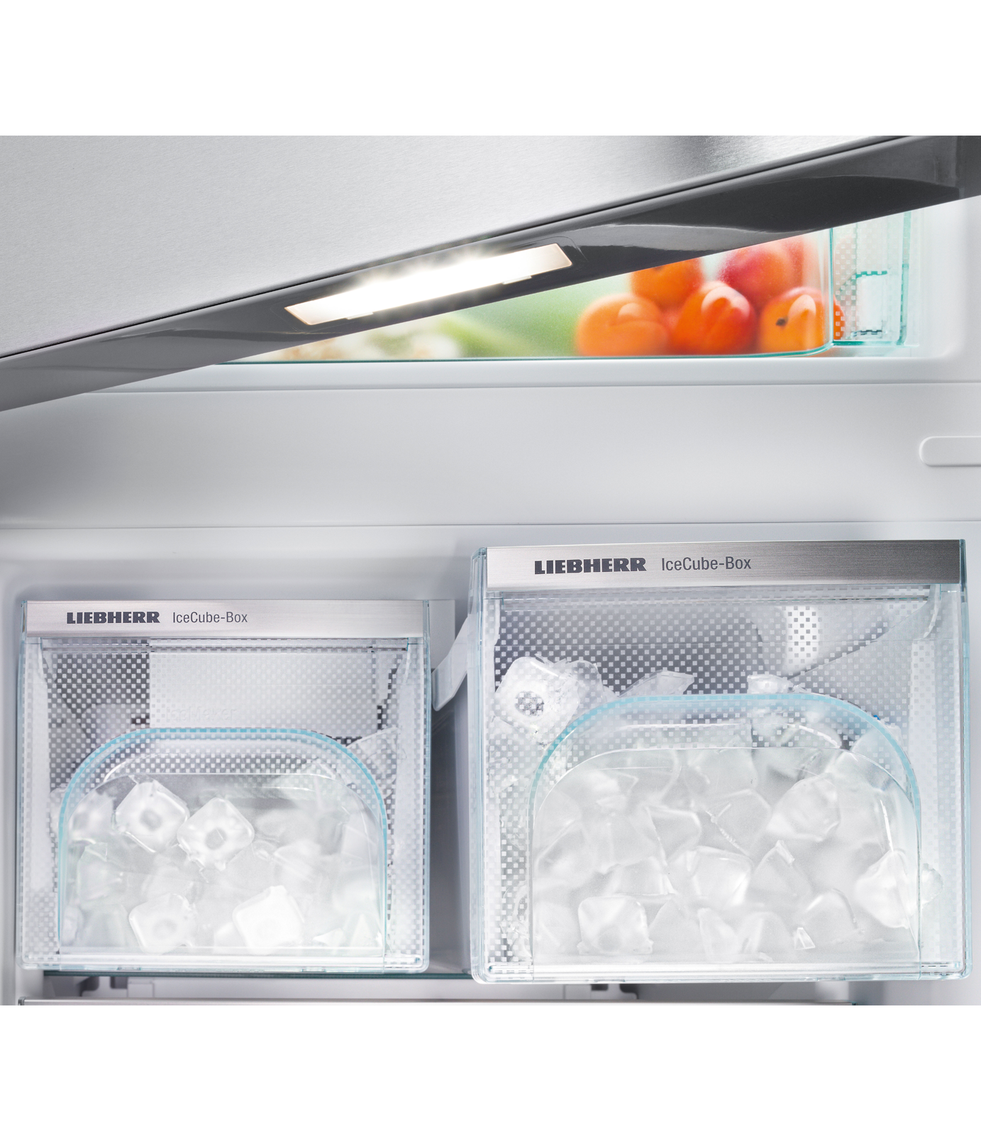 Inside a refrigerator, ice cube boxes are filled with ice. Apricots are visible in a compartment above. Bright overhead lighting.