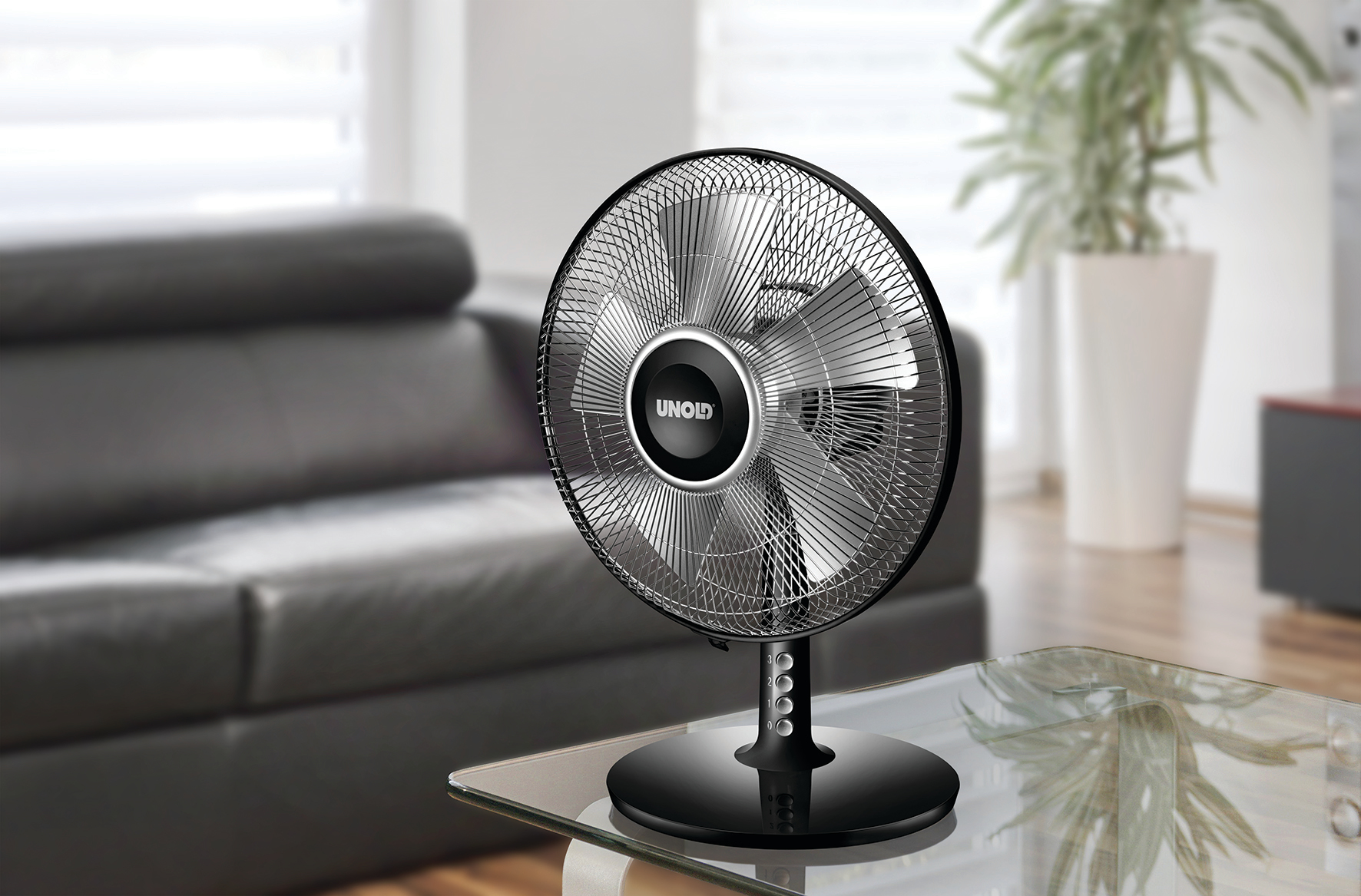 Schwarz-silberner Ventilator auf einem Glastisch, Sofa und Pflanze im Hintergrund.