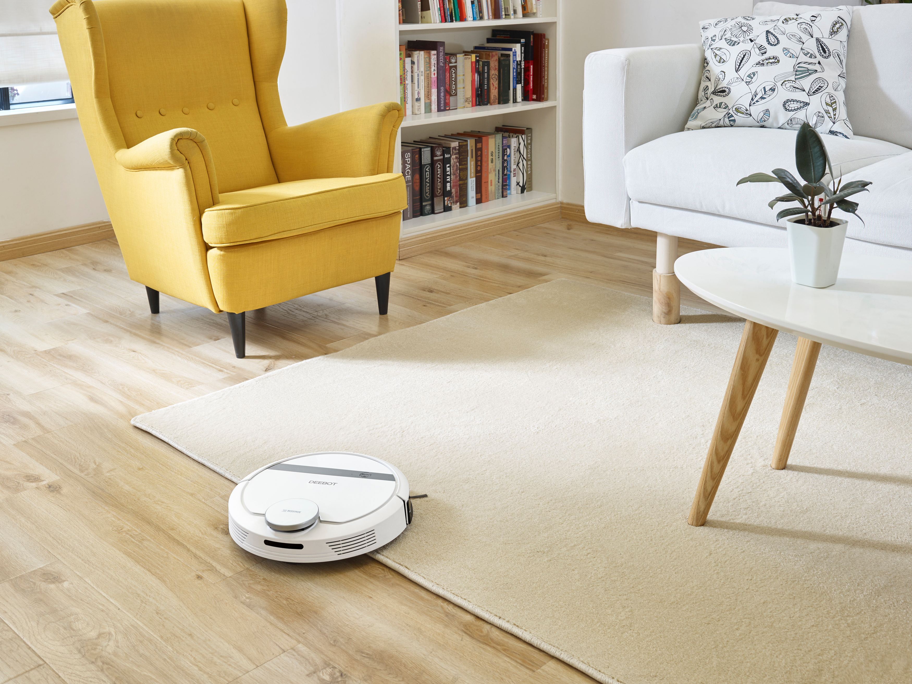 Ein weißer Roboter-Staubsauger reinigt ein Wohnzimmer. Ein gelber Sessel, eine weiße Couch und ein runder Tisch sind im Raum.