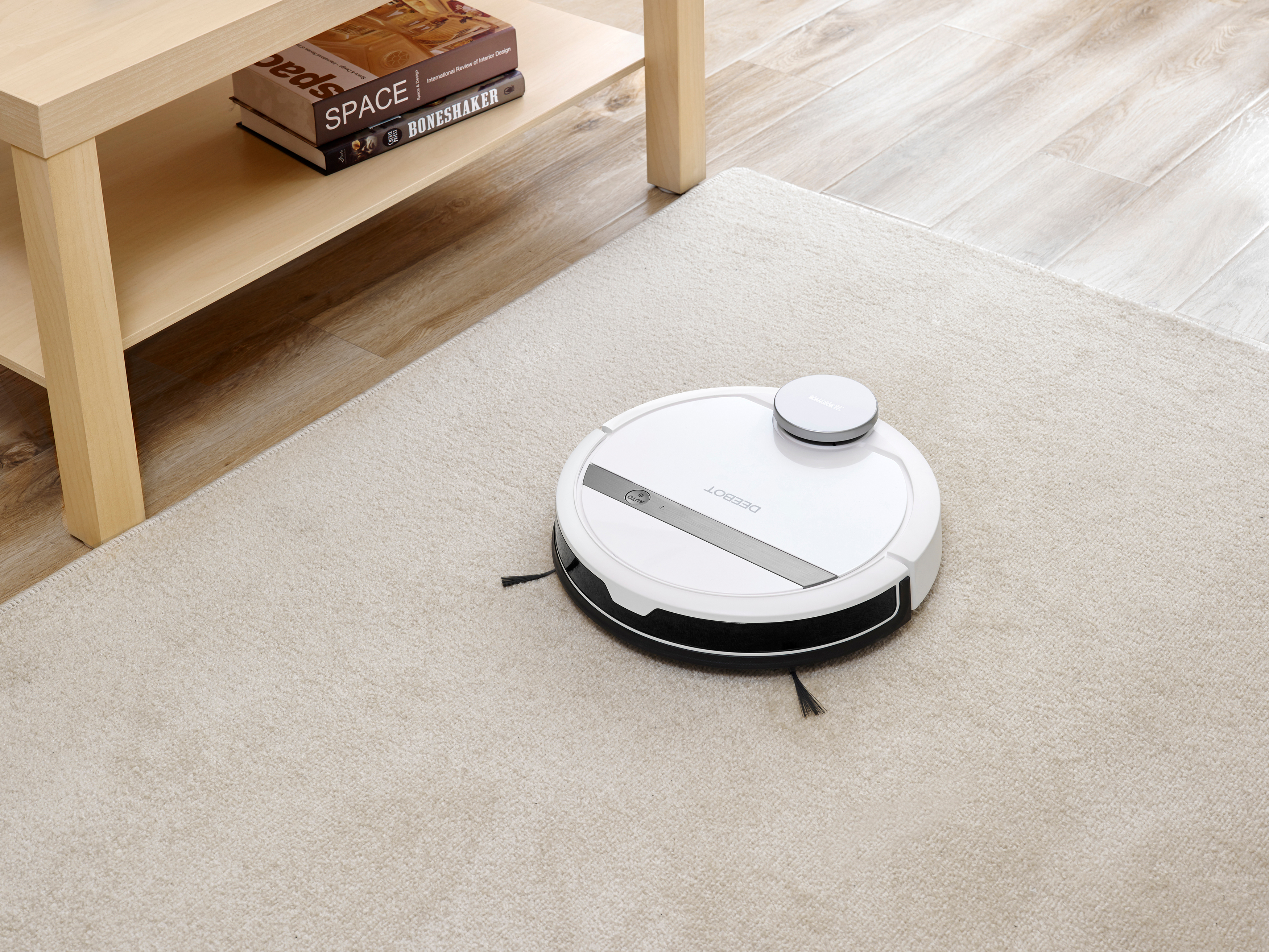Weißer Roboter-Staubsauger auf beigem Teppich. Ein Holztisch mit Büchern links. Holzboden im Hintergrund.