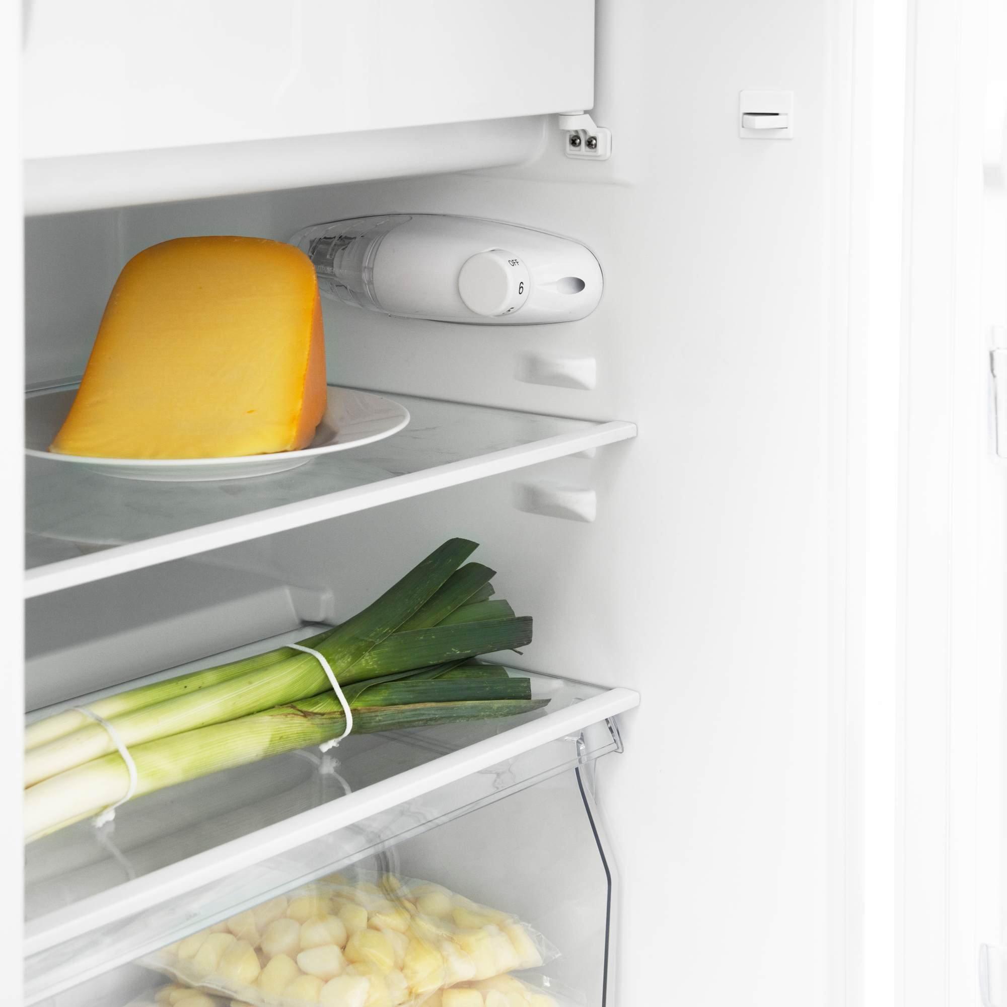 A wedge of cheese on a plate and leeks in a refrigerator.