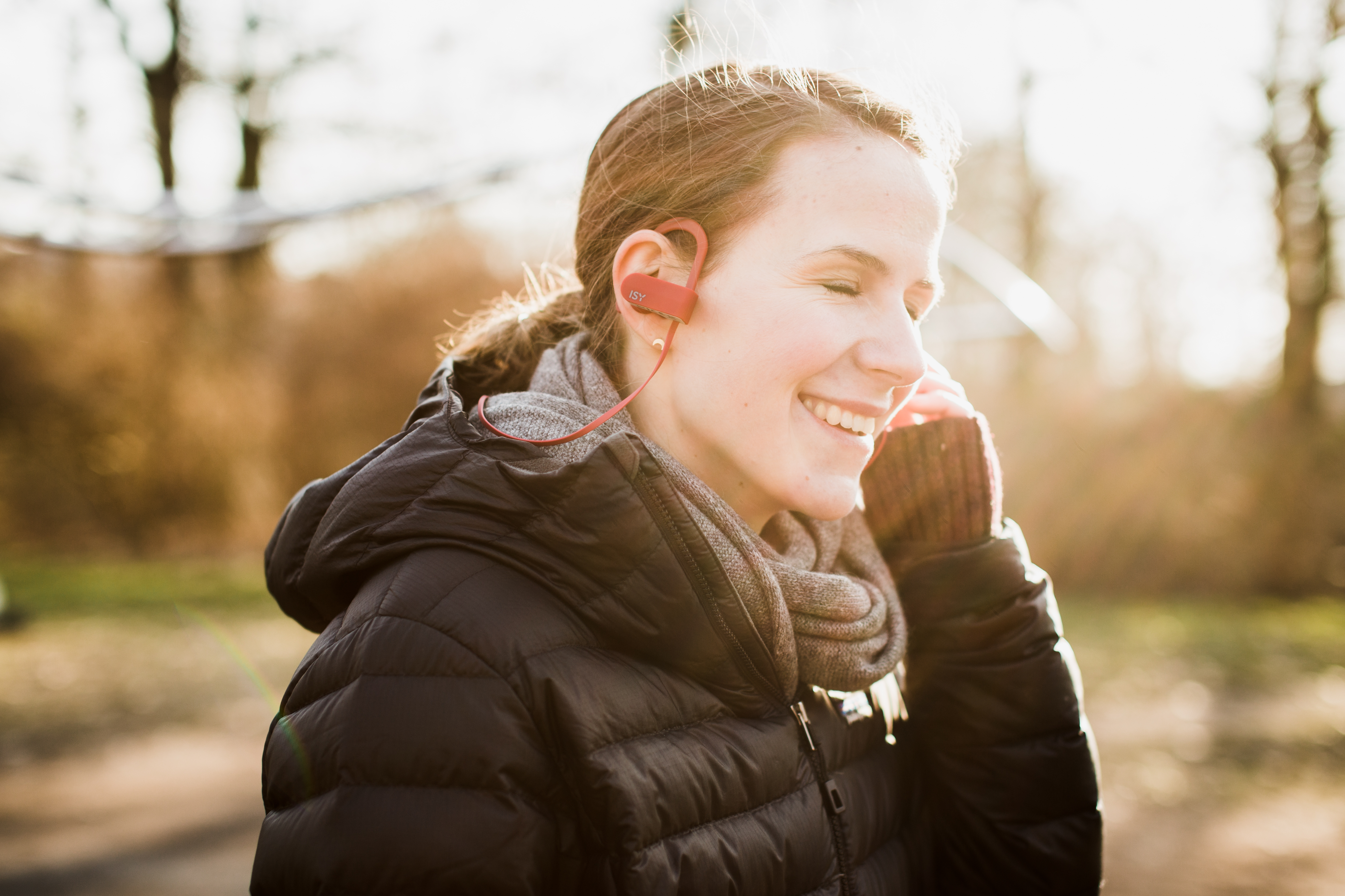 Eine Frau mit roten Ohrhörern lächelt draußen, trägt eine schwarze Jacke und einen Schal.