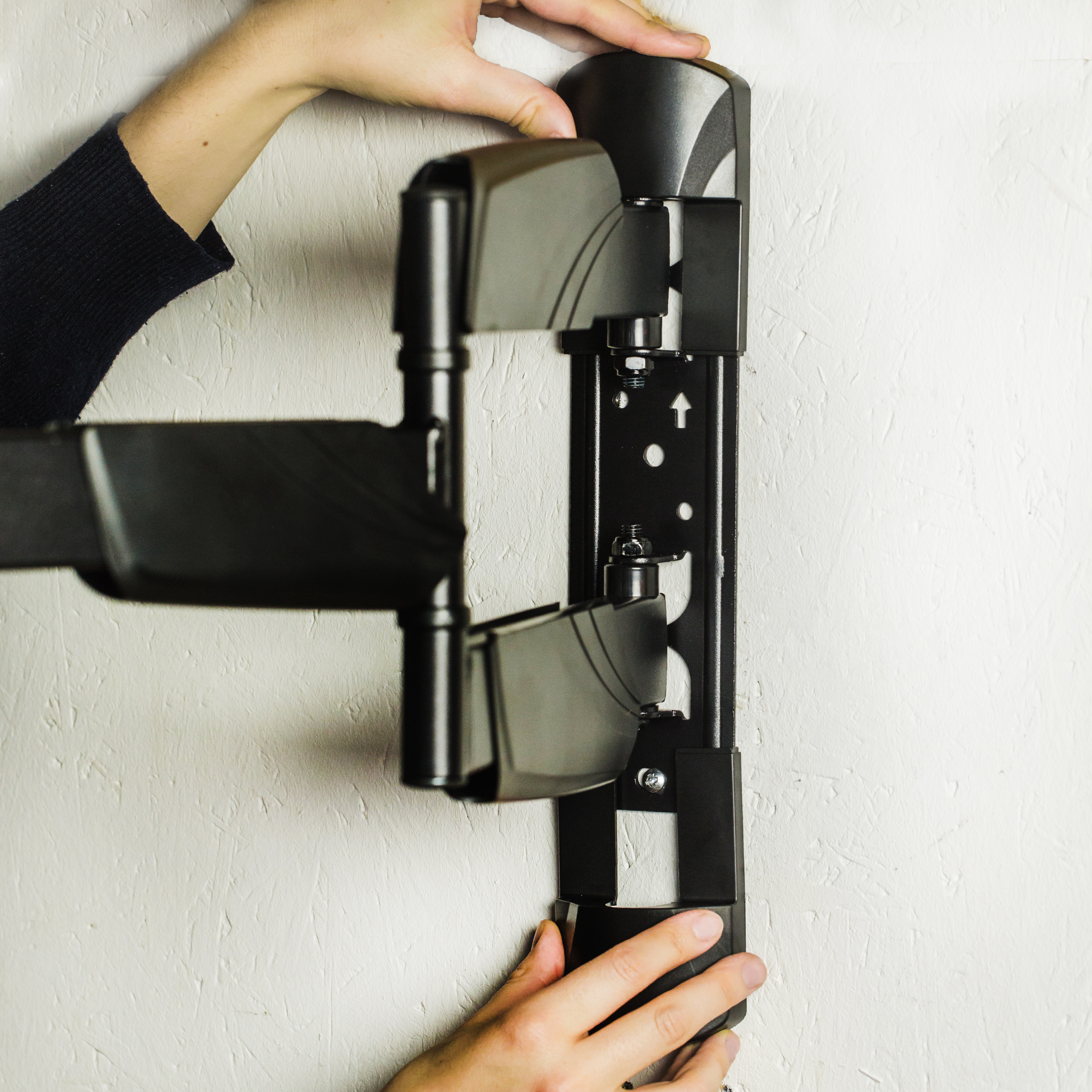 Mains installant un support mural de télévision noir sur un mur blanc texturé.