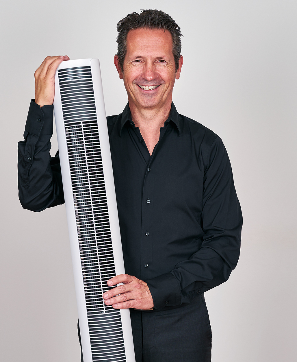 Ein Mann in schwarzem Hemd hält einen weißen Turmventilator und lächelt in die Kamera. Der Hintergrund ist schlicht.