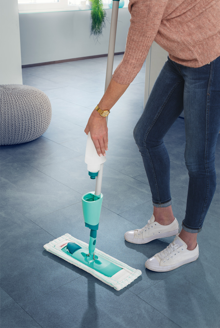 Eine Person, die einen Sprühwischer auf einem blauen Boden verwendet, trägt weiße Turnschuhe und Jeans.