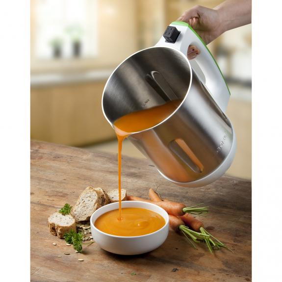 Un appareil à soupe argenté versant de la soupe orange dans un bol blanc sur une table en bois.