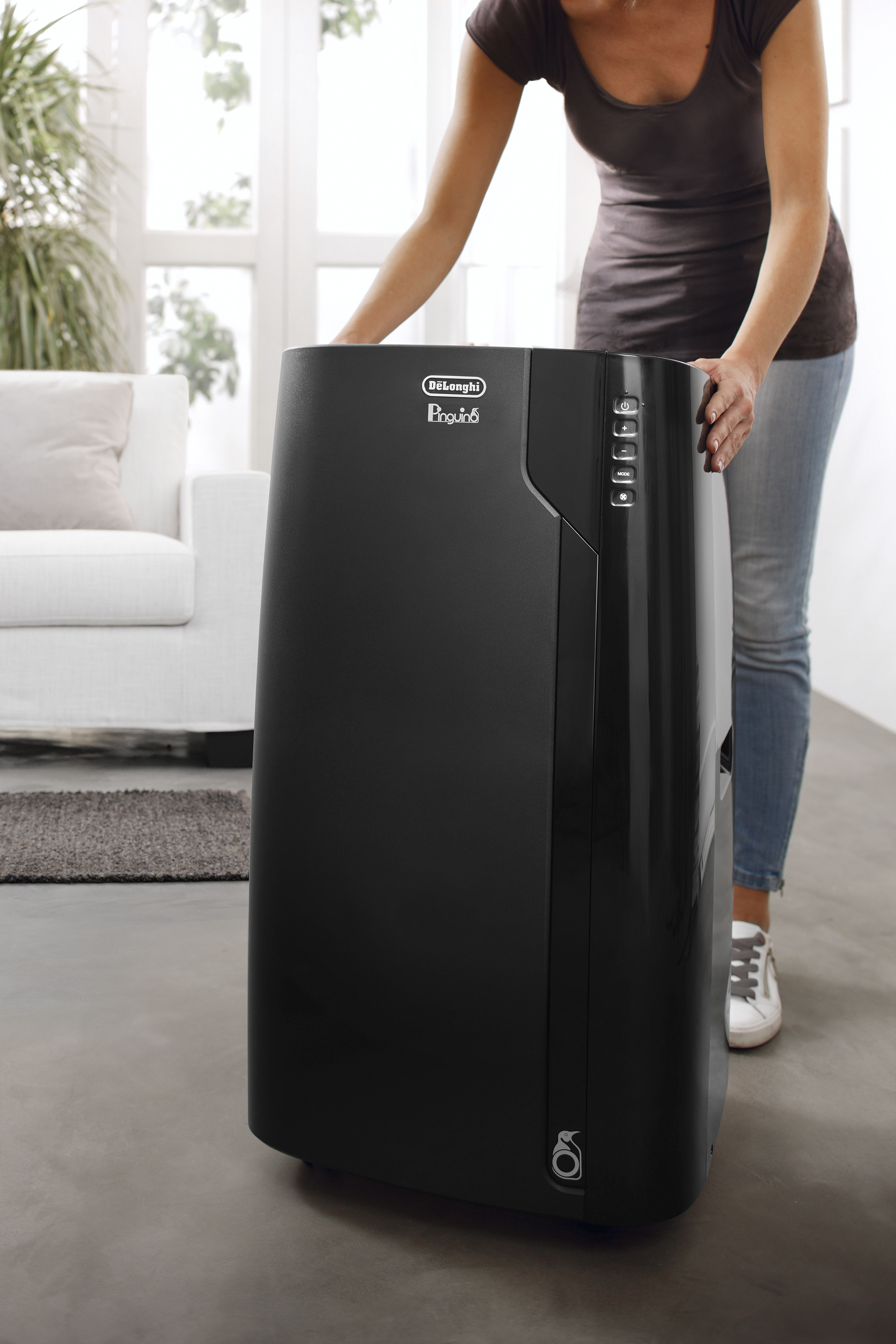 Een vrouw past een zwarte DeLonghi airconditioner aan. Een witte bank en een raam zijn op de achtergrond.