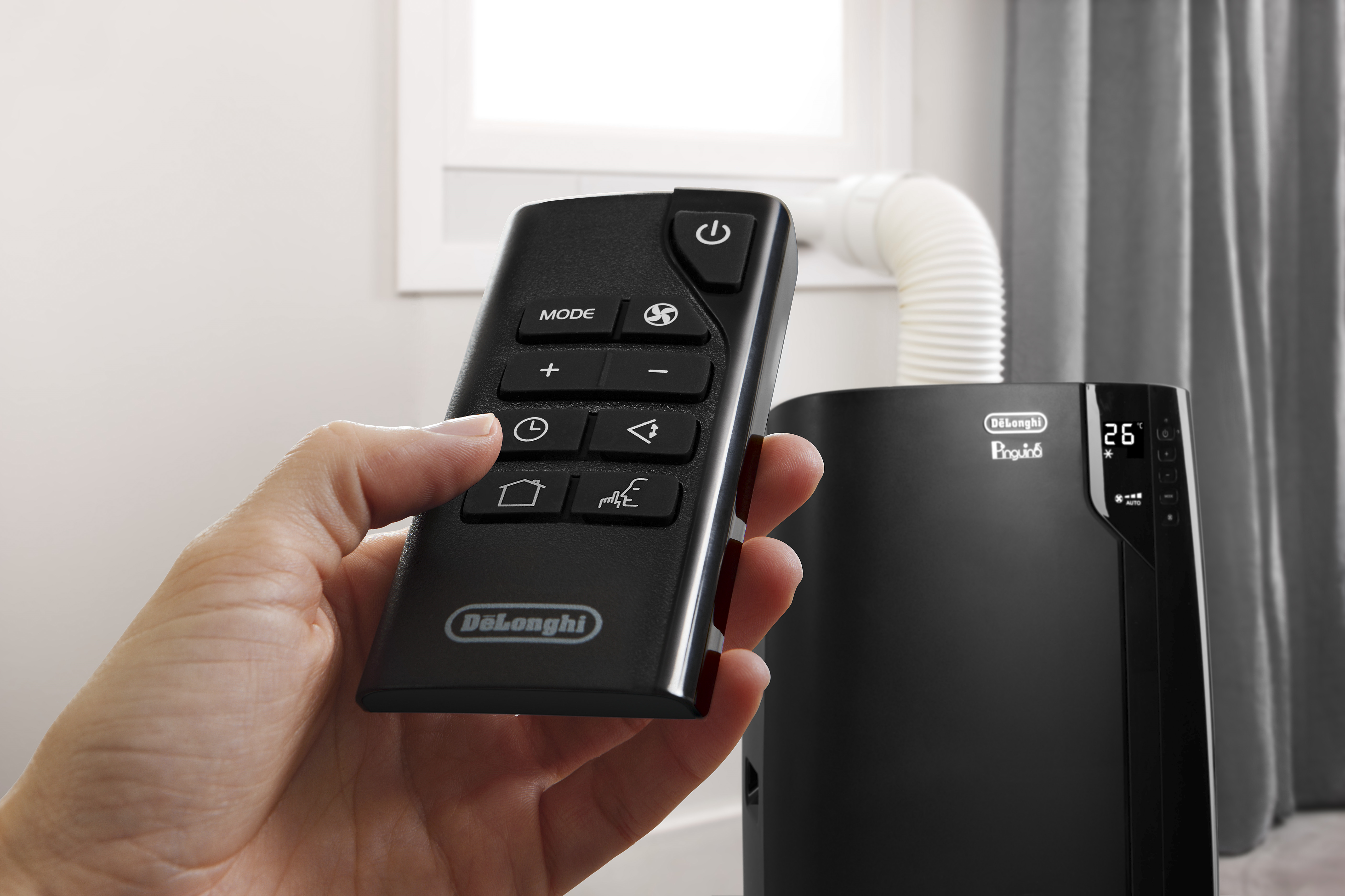 Een hand houdt een zwarte afstandsbediening voor een DeLonghi airconditioner vast. Een zwarte airconditioner staat op de achtergrond met een witte uitlaatslang.
