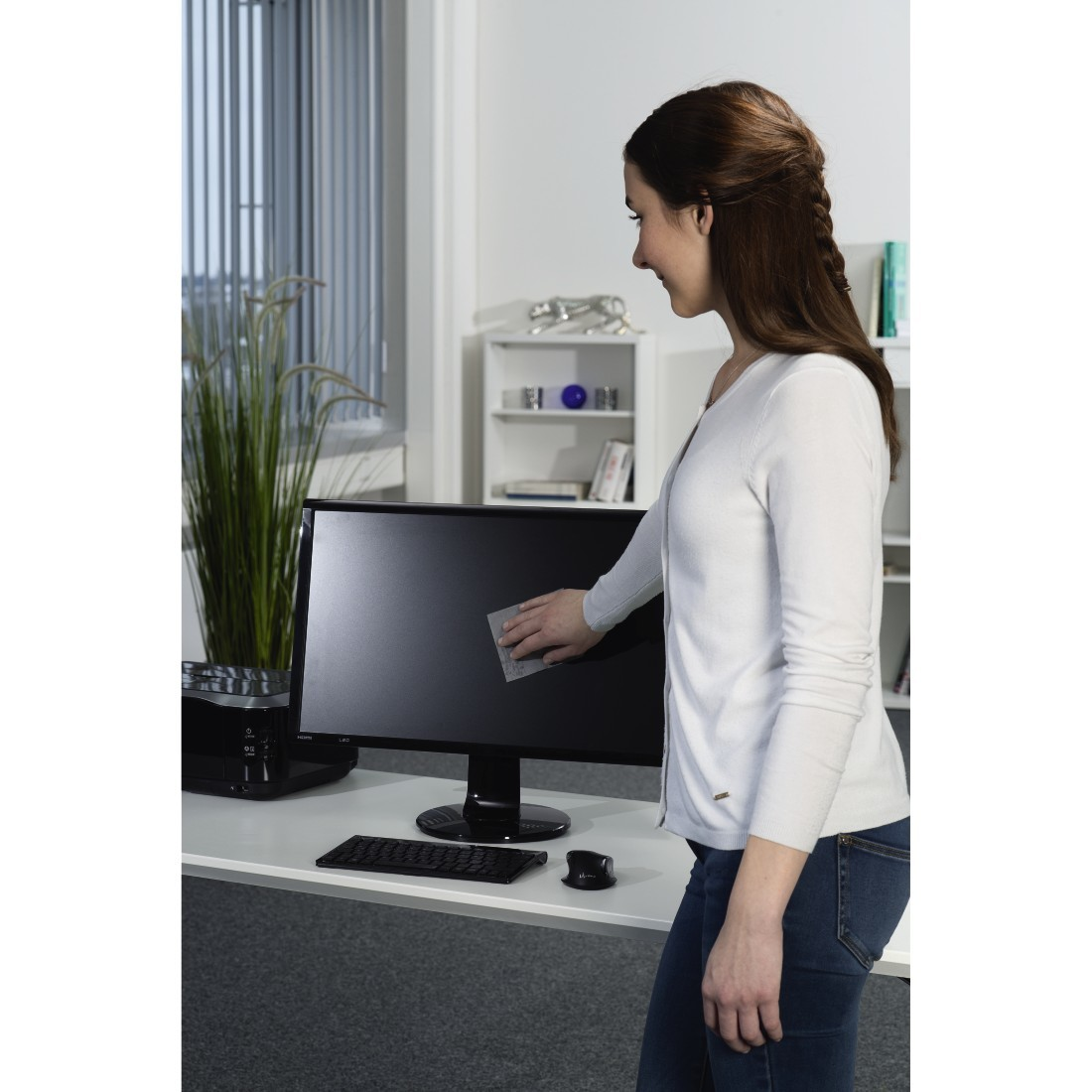 Une femme essuyant un écran d'ordinateur avec un chiffon de nettoyage sur un bureau blanc, avec une plante.