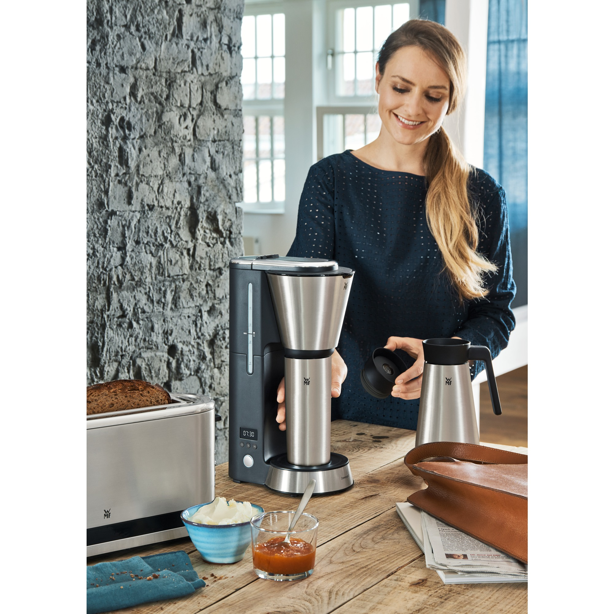 Vrouw die koffie zet met een koffiezetapparaat, een broodrooster en ontbijtproducten op tafel.
