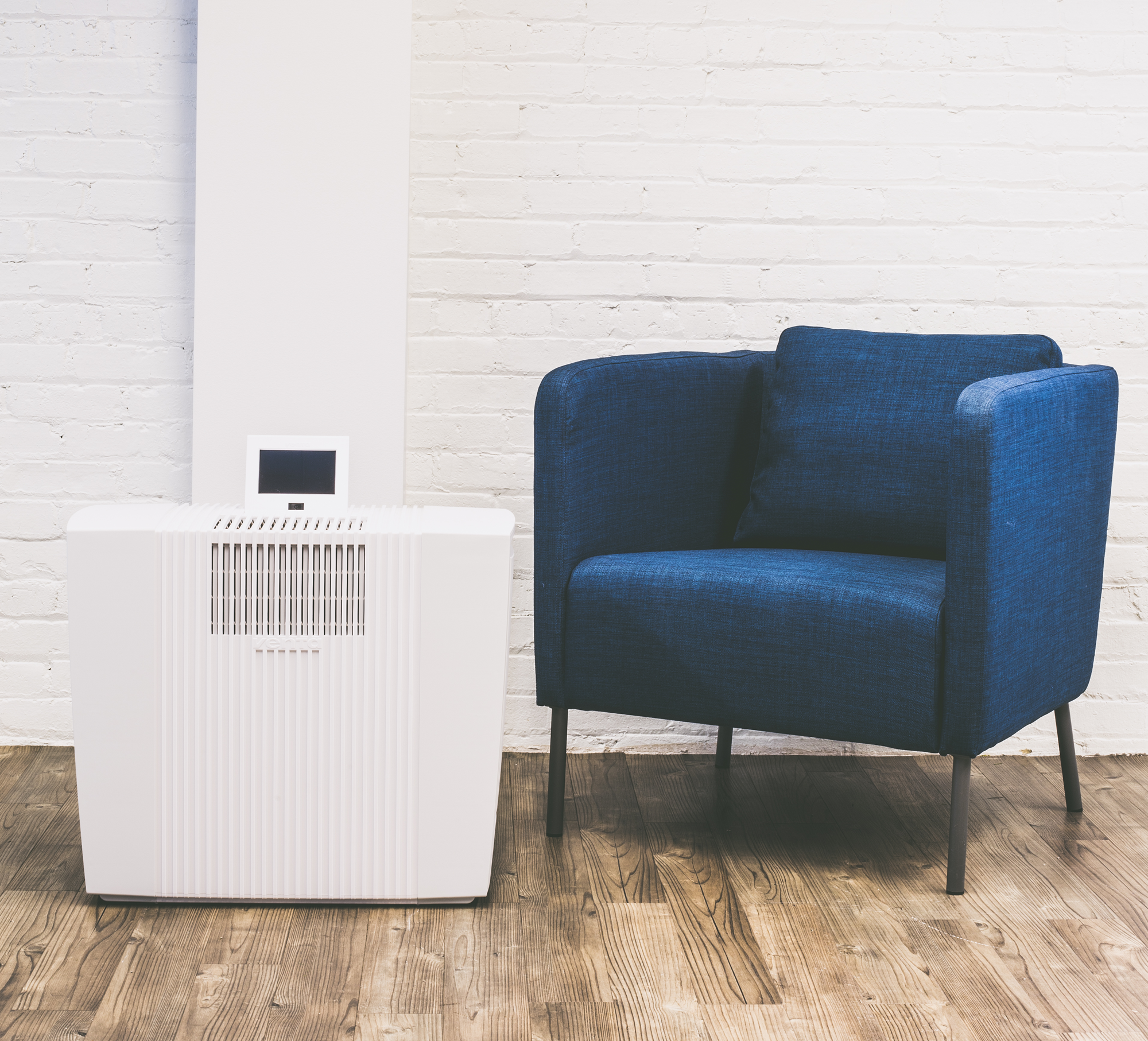 A white air purifier and blue armchair in a room with wooden floor and a white brick wall.