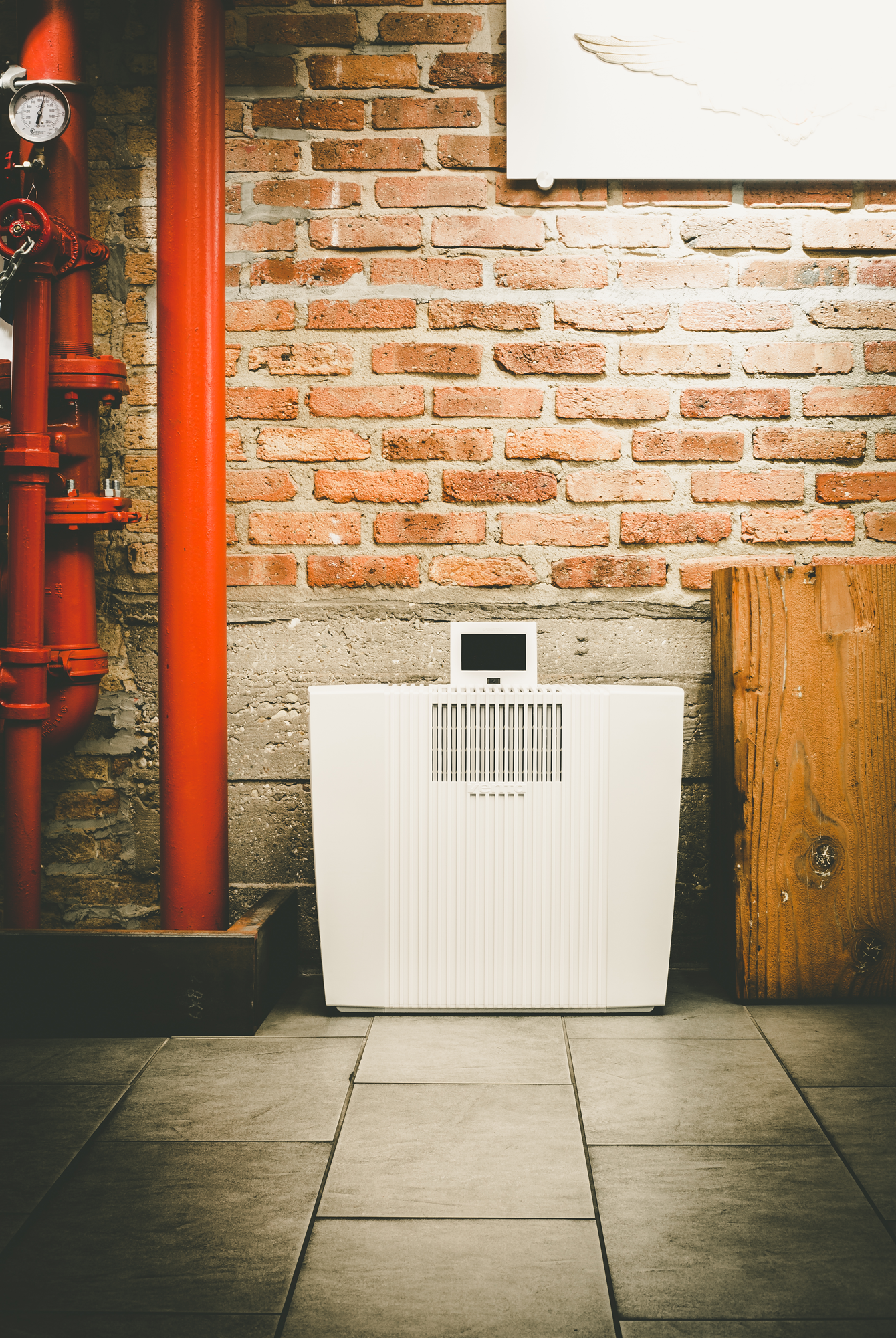 An industrial setting with a white air purifier, red pipes, brick wall, and concrete floor.