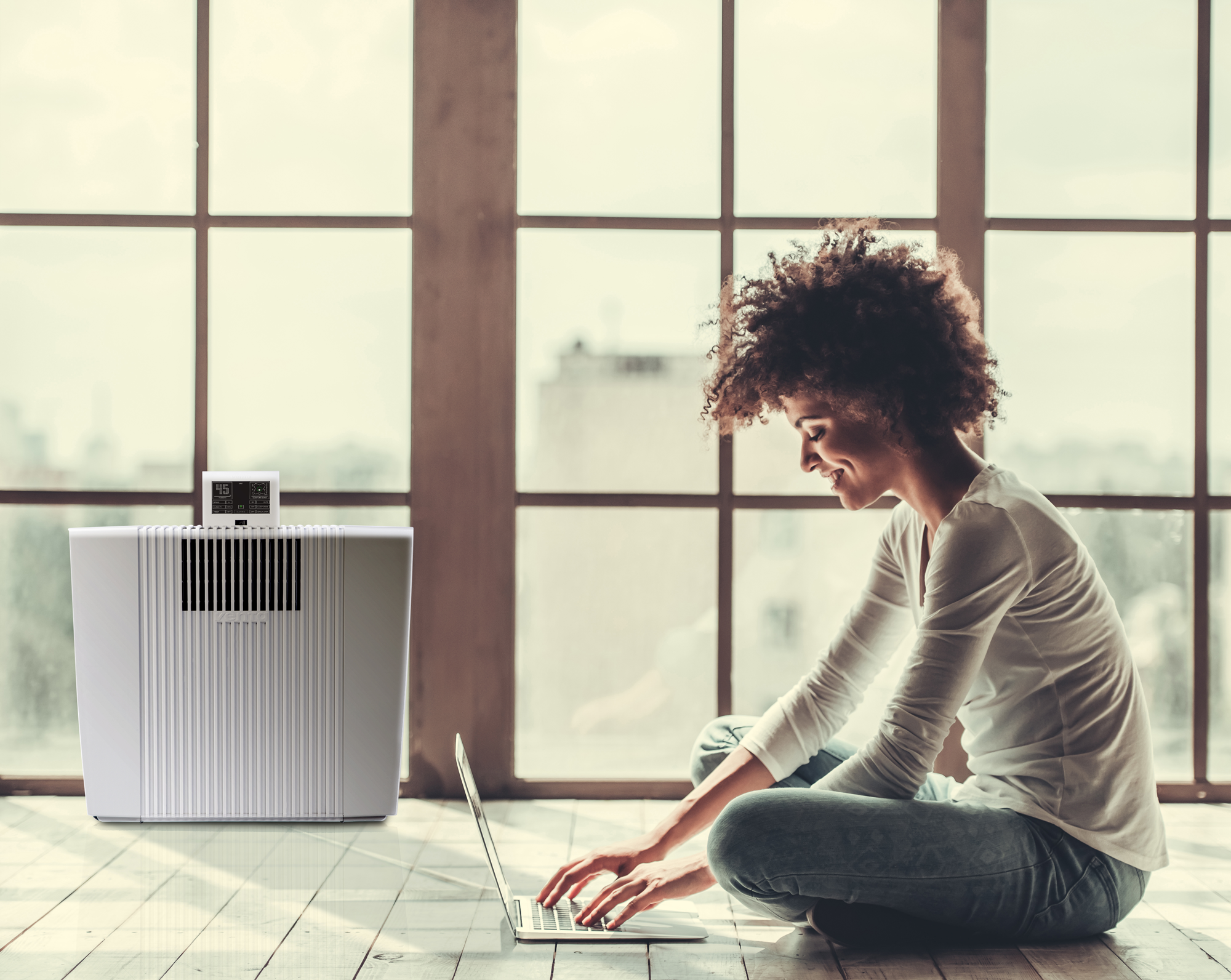Woman with laptop sits by a window, air purifier to the left.