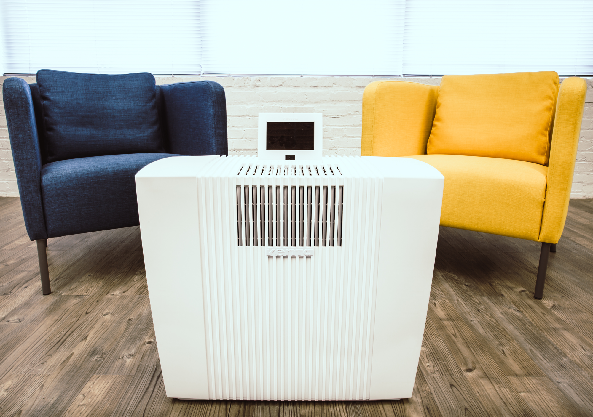A white air purifier between blue and yellow armchairs. Wooden floor and blinds in the background.