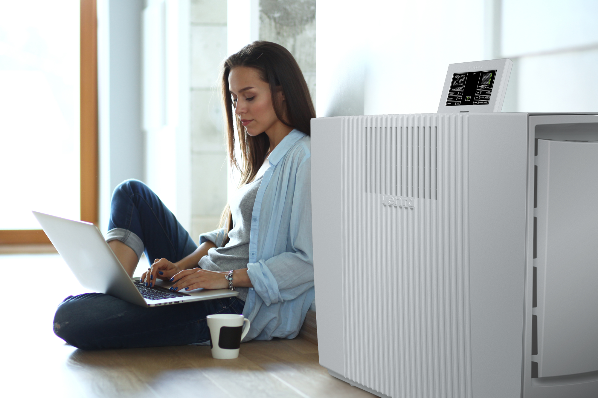Woman using laptop on floor next to a white air purifier and a mug.