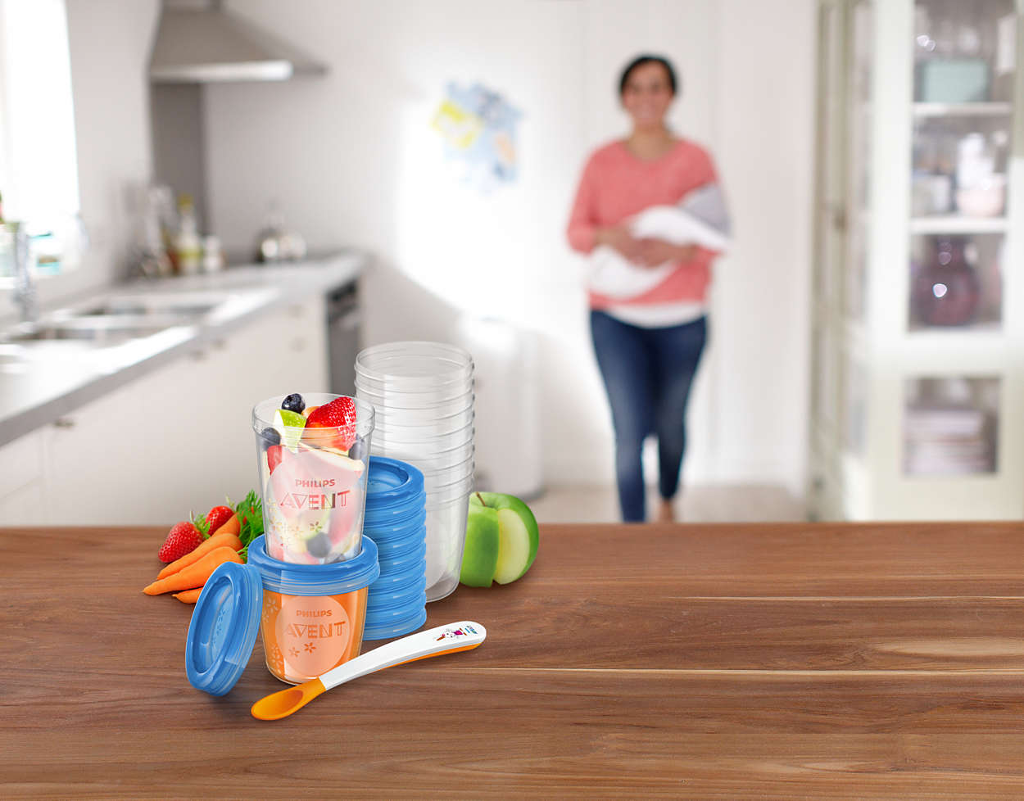 Bébiétel-tartók és kanál az asztalon. Egy nő babát tart a konyha hátterében.