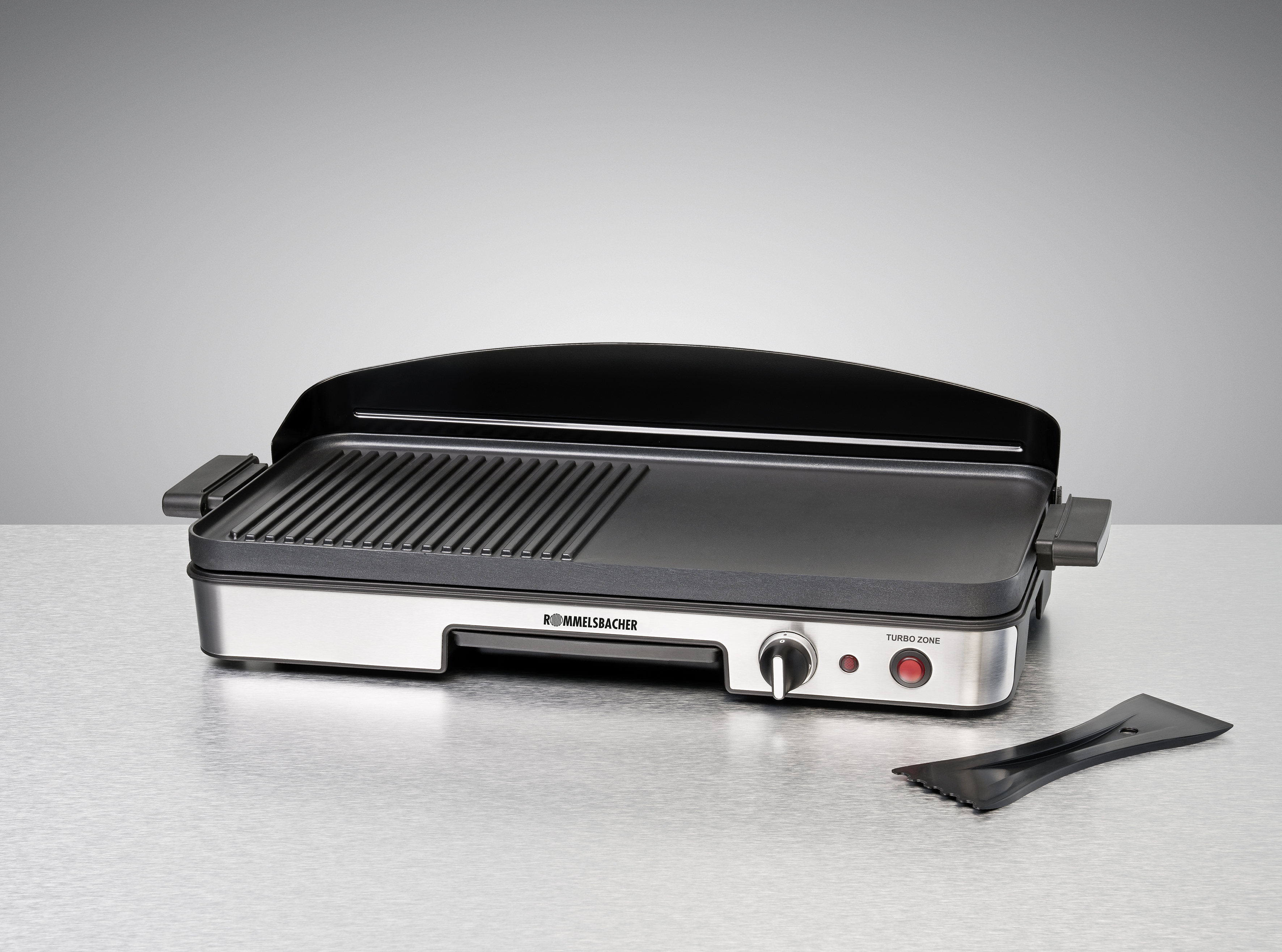 Elektrischer Grill mit schwarzer Oberfläche und silbernem Rahmen, mit Spatel daneben.