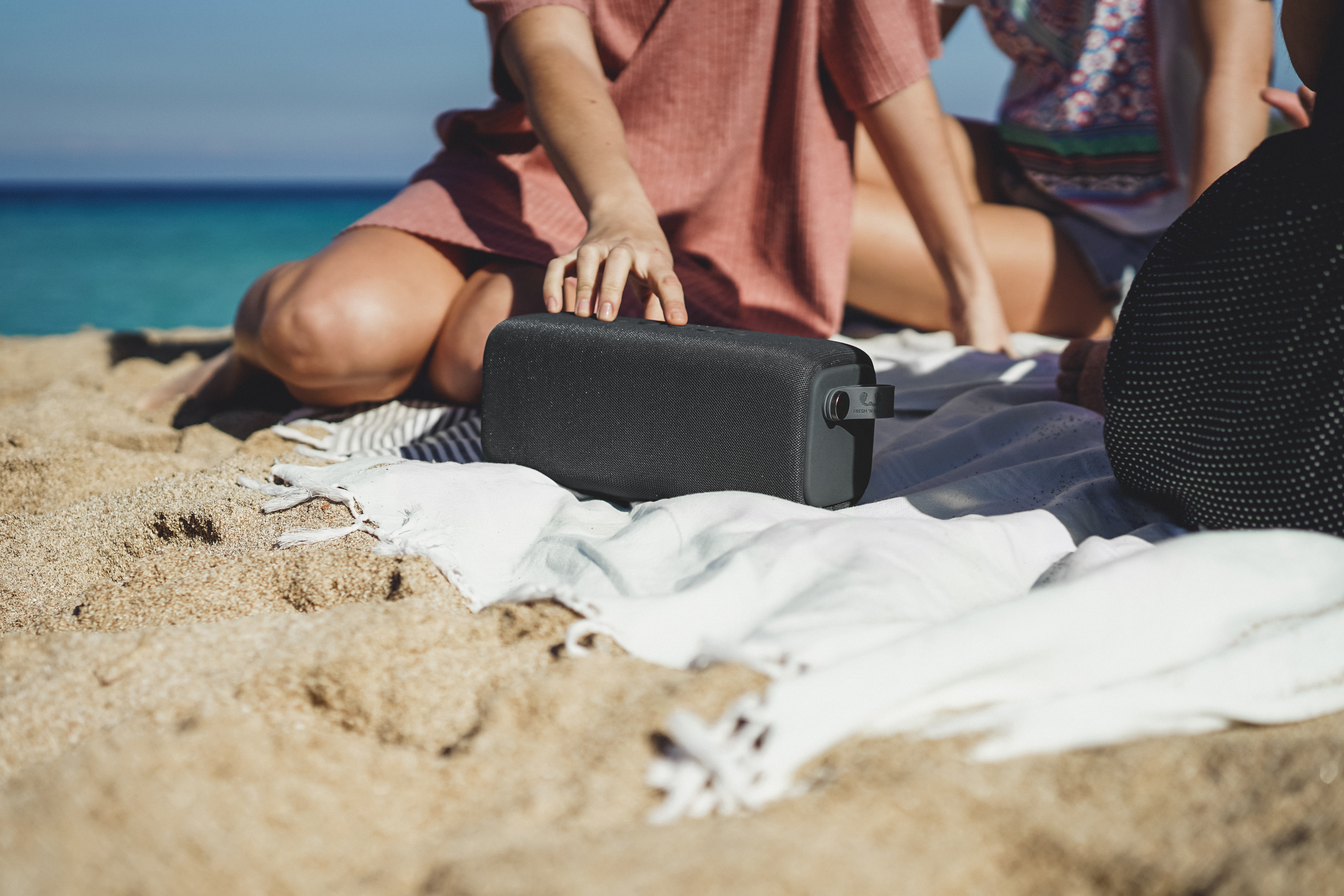 Zwei Leute am Strand, einer hält einen schwarzen Lautsprecher auf einer Decke, Meer im Hintergrund.