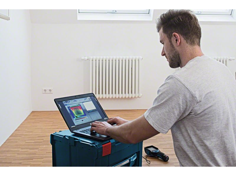 Férfi laptopot használ egy kék szerszámosládán, fehér falú szobában radiátorral.