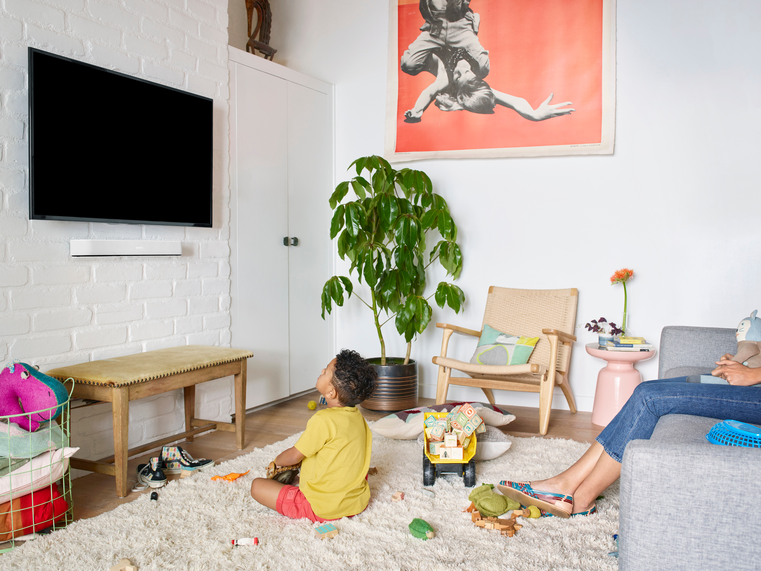 Wohnzimmer mit Fernseher, spielendem Kind und Poster. Möbel und eine große Pflanze sind vorhanden.